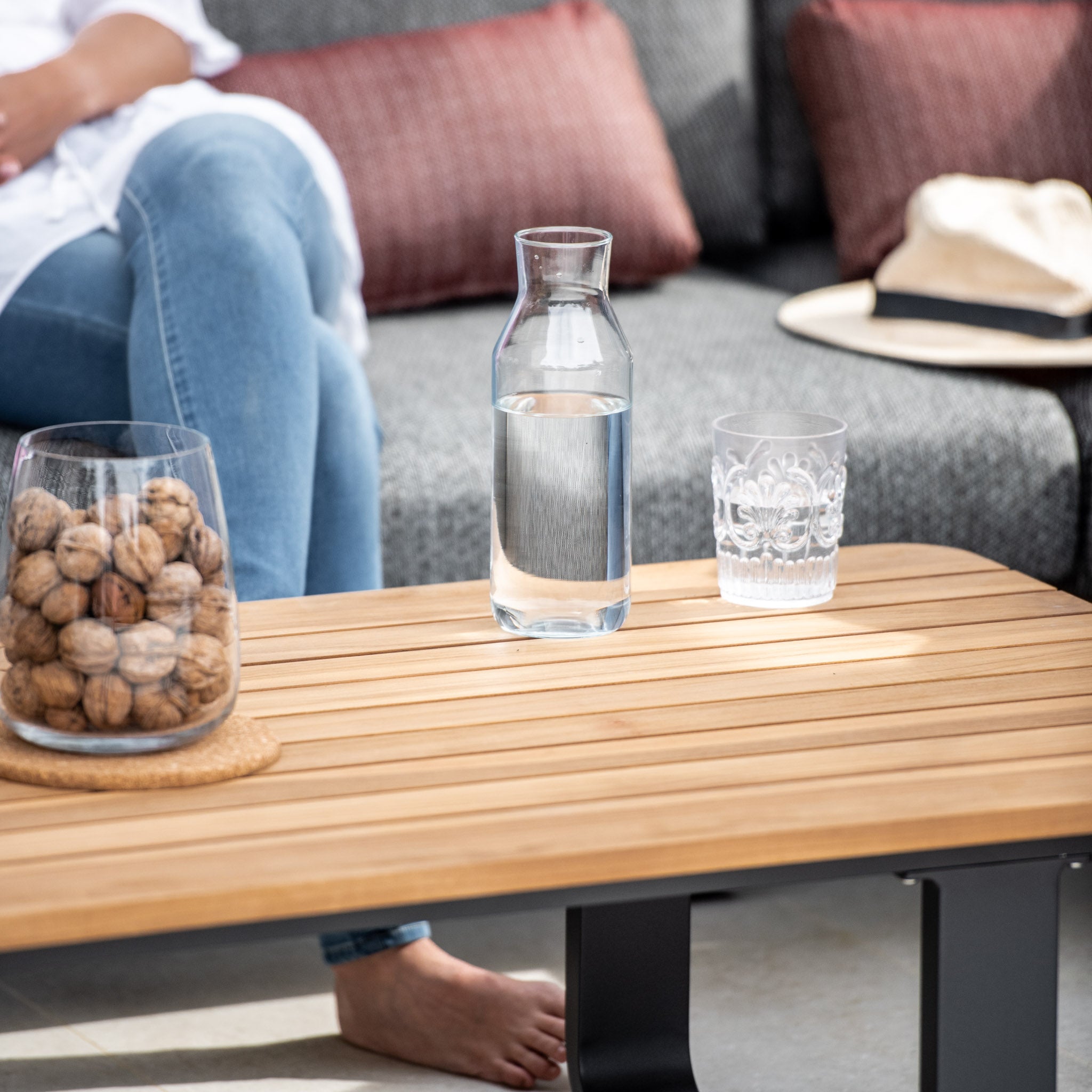 A wooden teak coffee table with a glass of water, a carafe, and a glass jar of walnuts sits before an Olive Green Bay Corner Group Set with 4 waist pillows; a person is seated in the background.