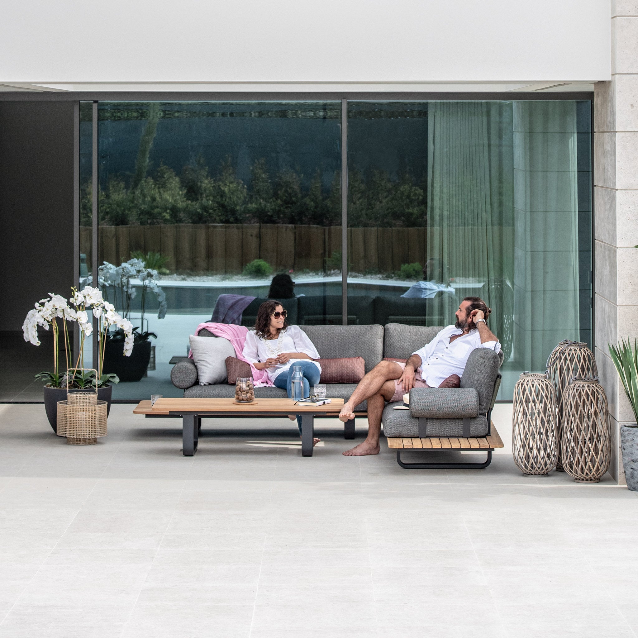 Two people relax on a Bay Corner Group Set with 4 Waist Pillows Teak Table in Olive Green in front of glass doors, surrounded by plants and modern decor, with an olive green acrylic rope accent adding a touch of elegance to the scene.