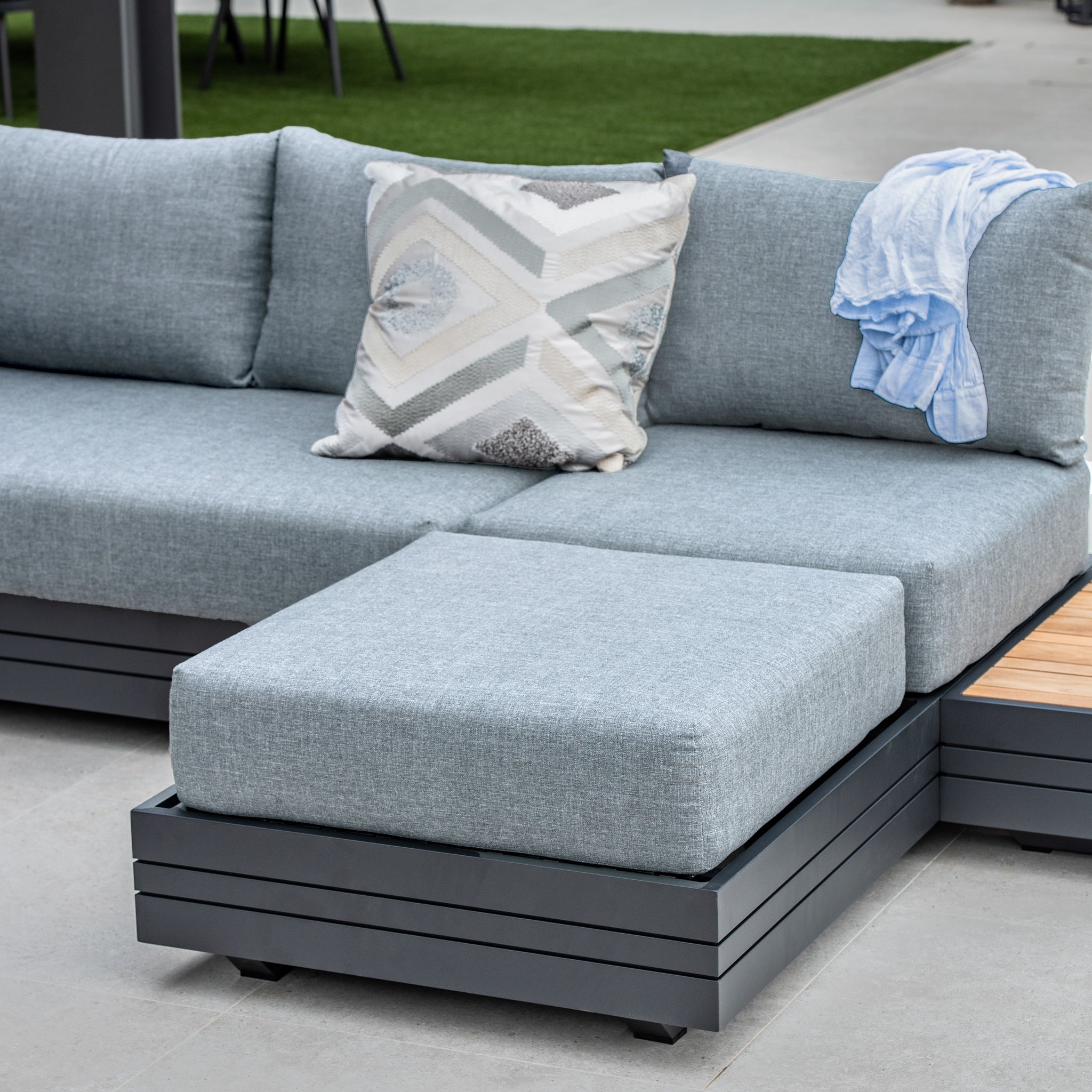 Experience the flexibility of our Panama Corner Unit/Footstool in Washed Grey, featuring a stylish gray outdoor sectional sofa adorned with a geometric patterned pillow and complemented by a blue cloth draped over the armrest.
