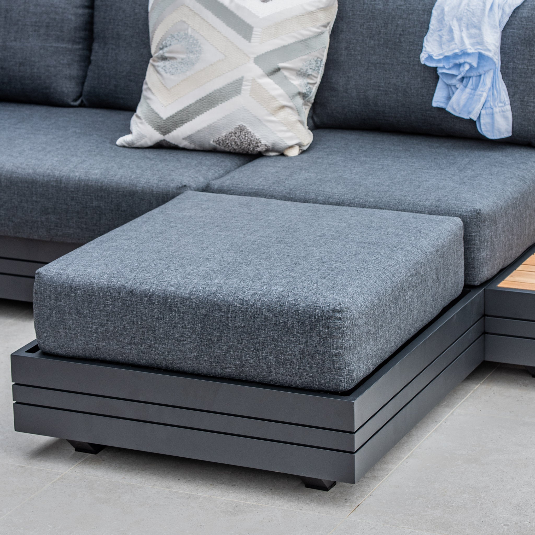 Panama Corner Unit/Footstool in charcoal with a patterned cushion, a blue shirt casually draped over the backrest, and matching footstool on the tiled floor.