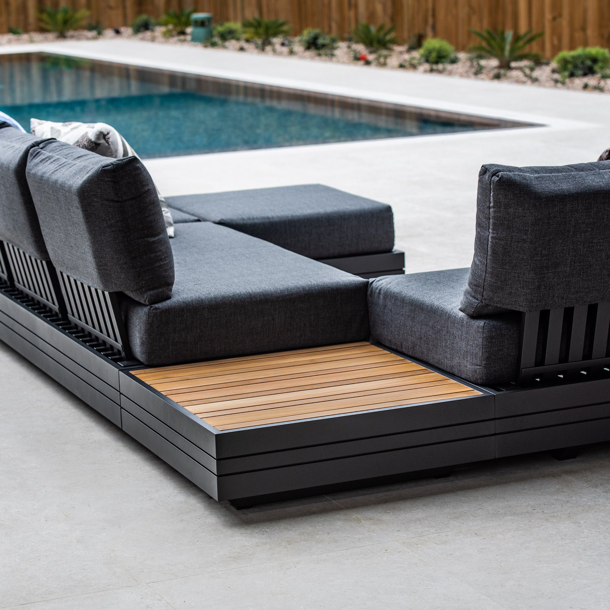 Modern outdoor lounge area by a pool, featuring dark gray cushioned seating and the luxurious Panama Teak Coffee Table with a teak wooden top, perfect for relaxing.