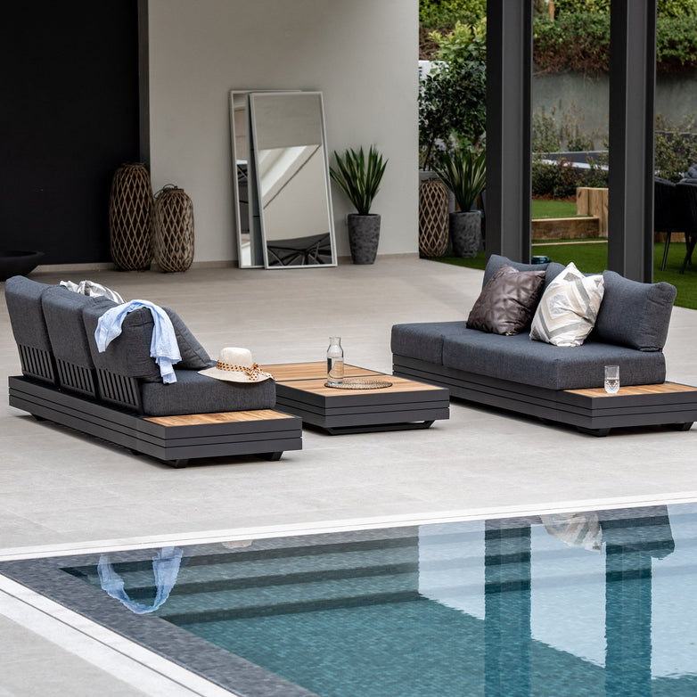 Modern outdoor patio with two gray cushioned loungers, a wooden coffee table, and a pool in the foreground, complemented by the sleek Panama 3 Seat Sofa with Sun Lounger Feature in Charcoal for added elegance.