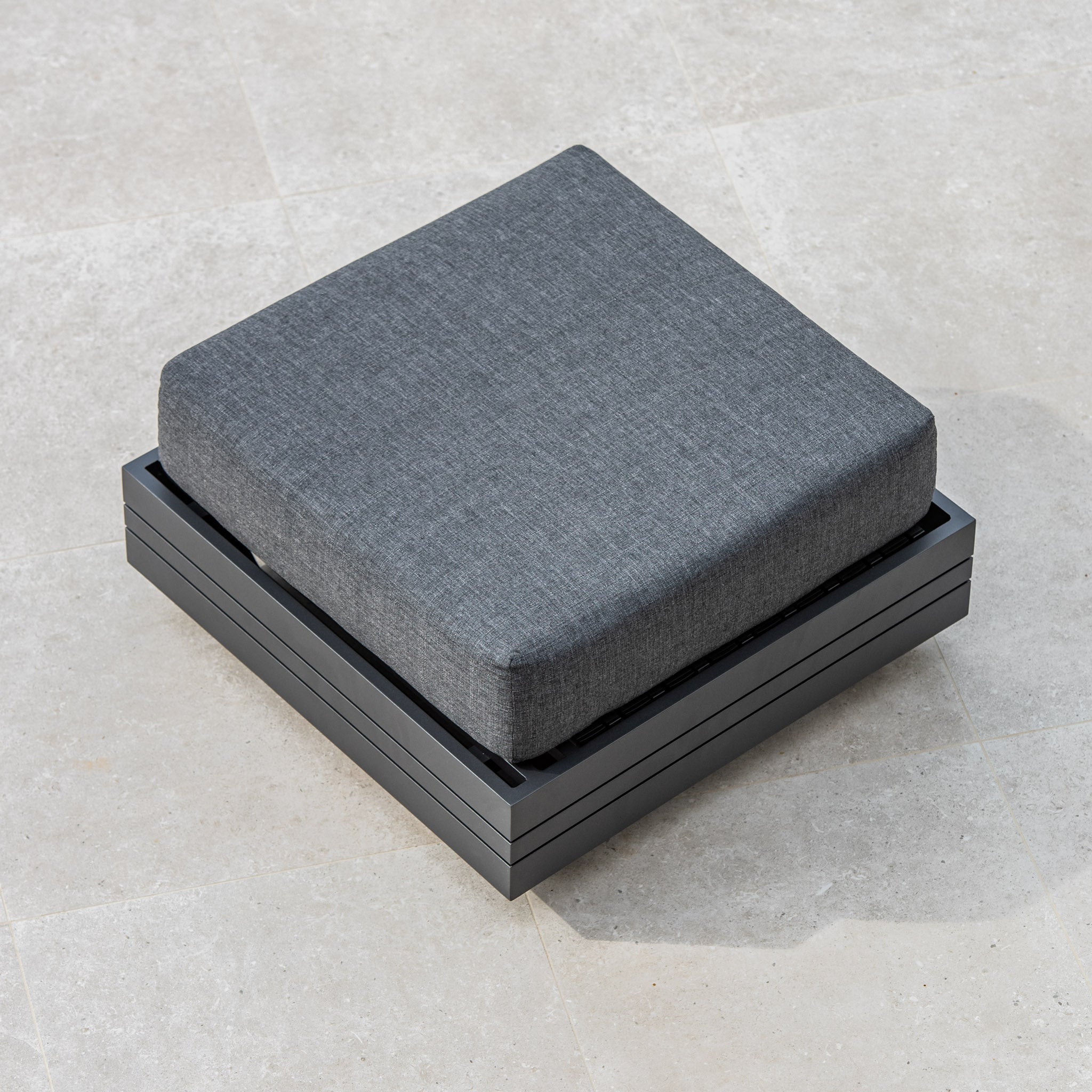 Top view of the Panama Corner Unit/Footstool in Charcoal, featuring a gray, square outdoor cushion on a matching dark gray metal base, placed on a light tiled floor. This versatile piece can function as part of a modular system or as a standalone corner unit.