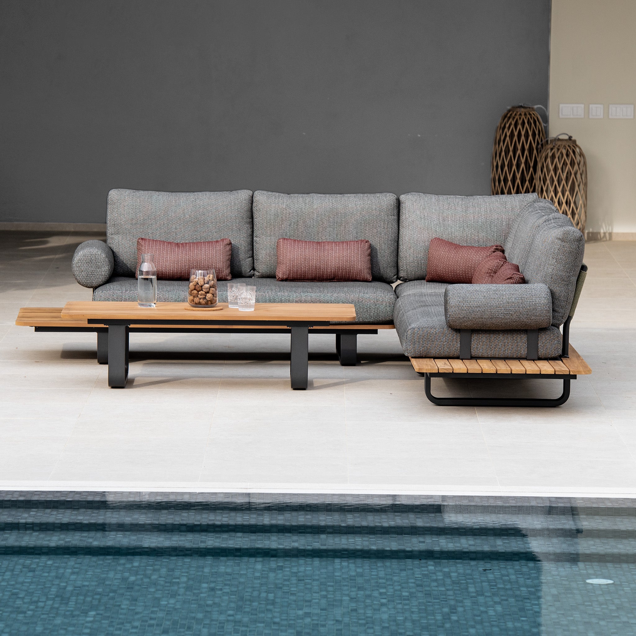 Modern outdoor patio with a Bay Corner Group Set, featuring 4 waist pillows and a teak table in olive green, along with drinks, snacks, and cushions near a pool.