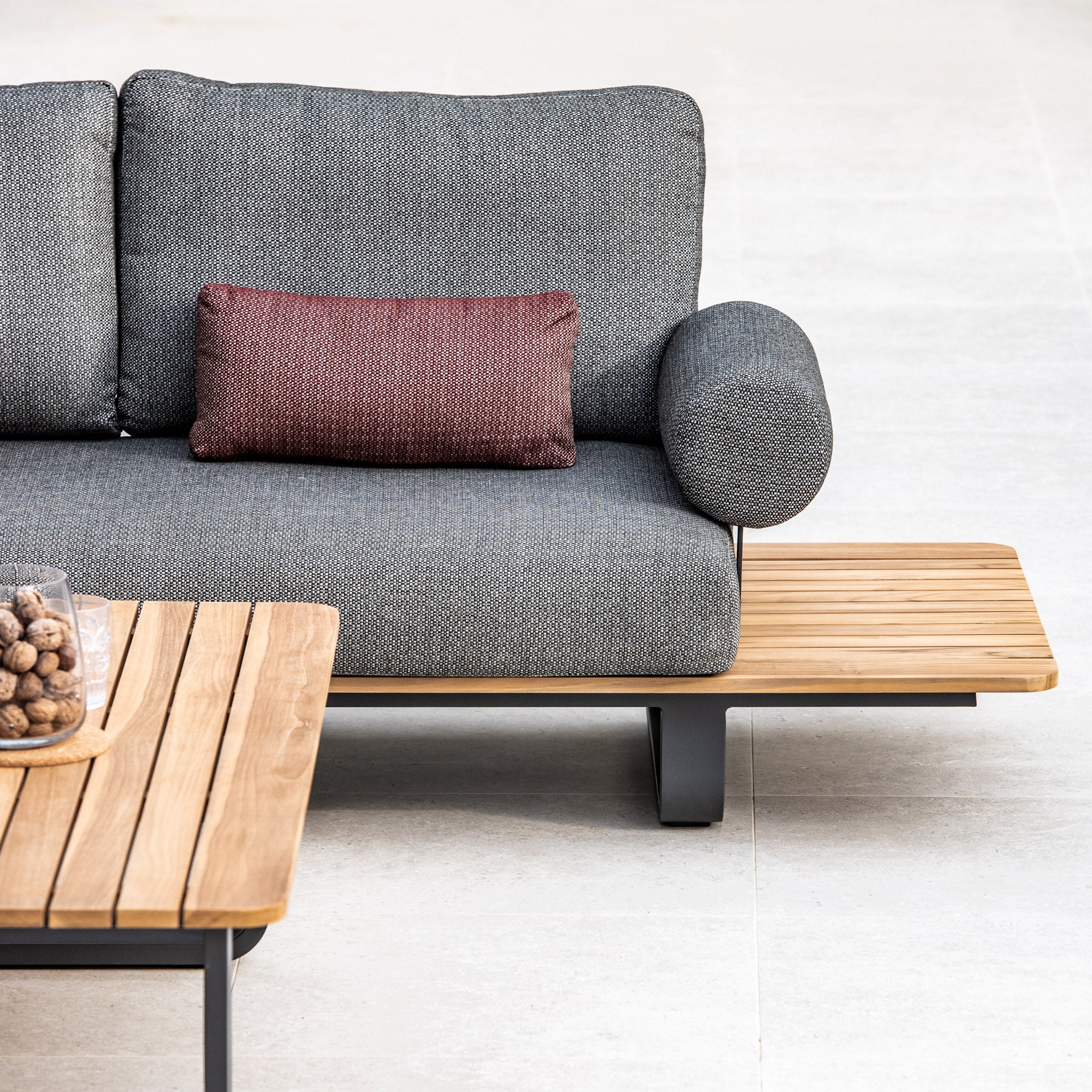 Bay Corner Group Set with gray cushions, red throw pillow, and adjacent teak side table. Teak coffee table in front, featuring stylish olive green acrylic rope accents and four waist pillows.