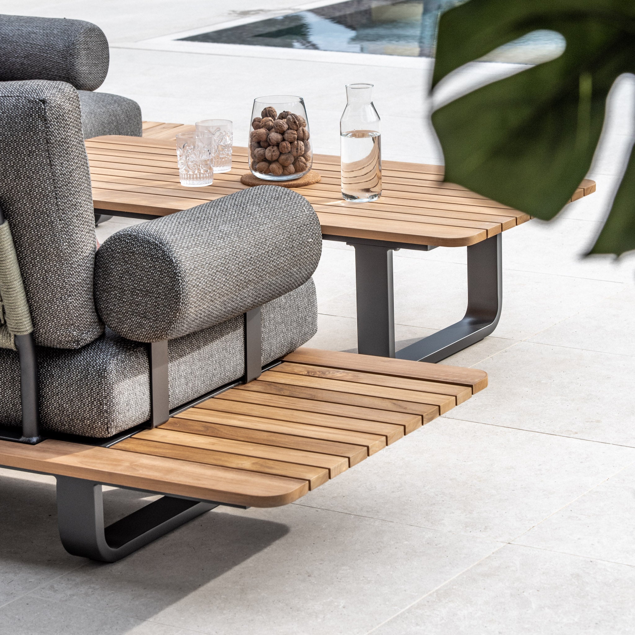 Modern outdoor seating area with the Bay Corner Group Set, featuring a sleek teak table, gray cushioned chairs with Olive Green acrylic rope detailing, four waist pillows, a glass pitcher, and a bowl of decorative items.