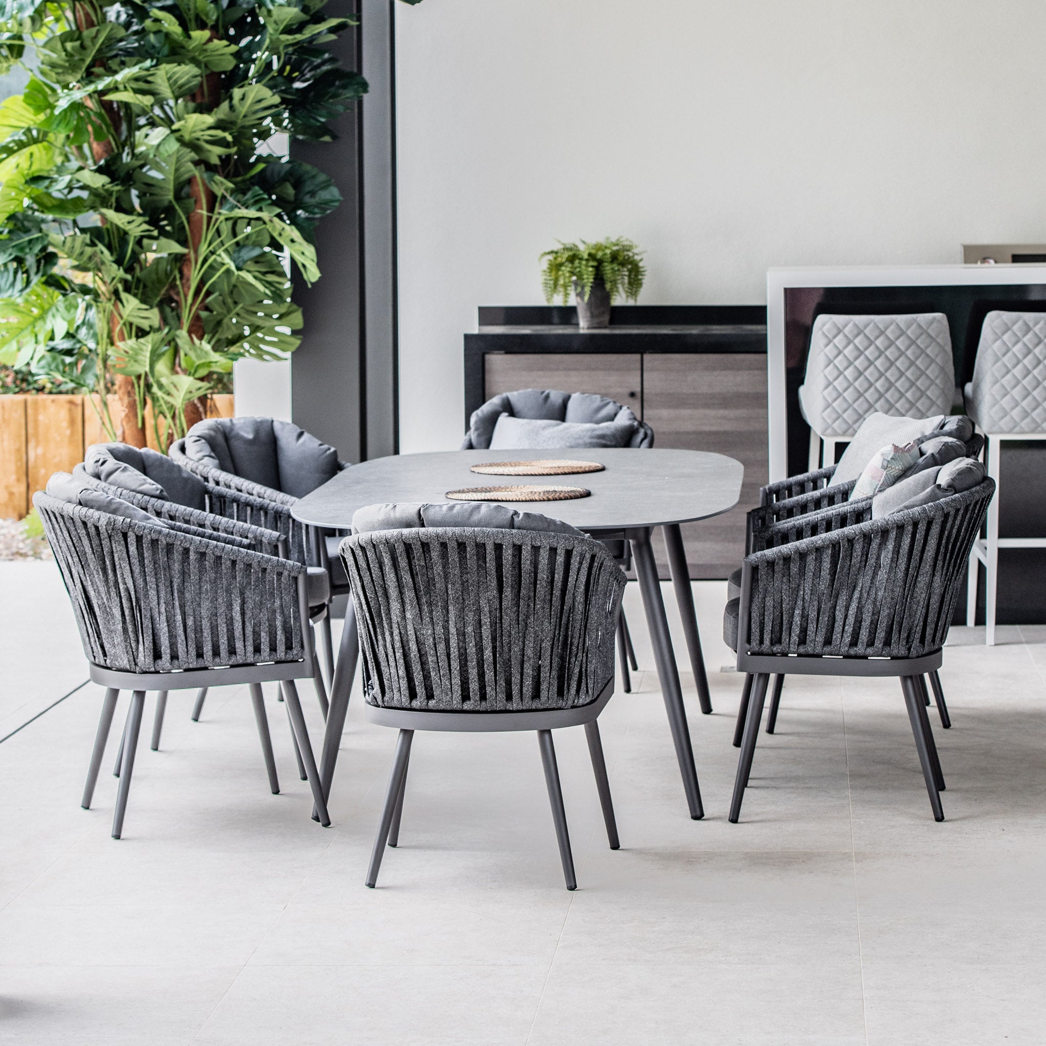 Modern outdoor patio featuring a Monterrey 8 Seat Rope Dining Set with an Oval Ceramic Table in Grey, a counter with three stools, and lush green plants in the background.
