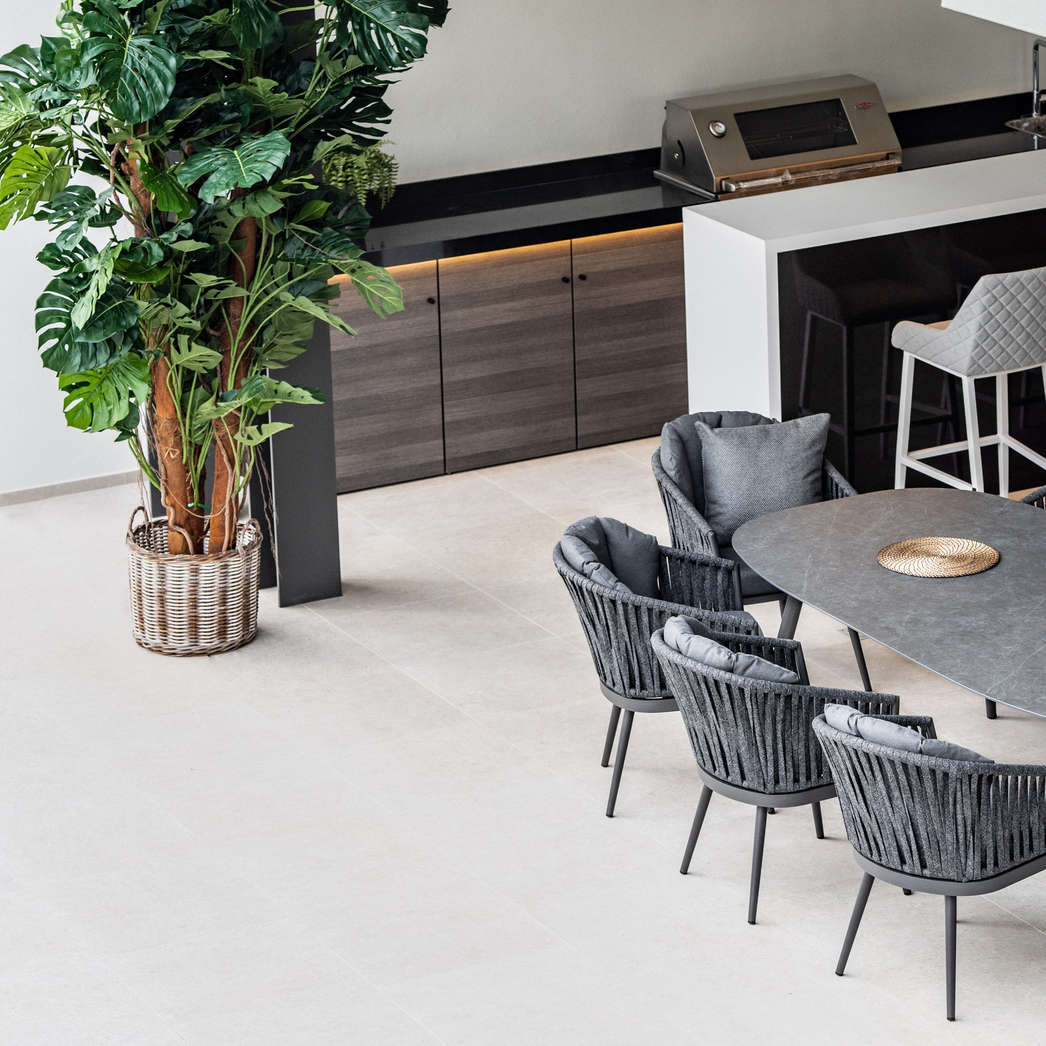 Modern indoor dining area featuring the Monterrey 8 Seat Rope Dining Set with Oval Ceramic Table in Grey, potted plants, and a grill station in the background.