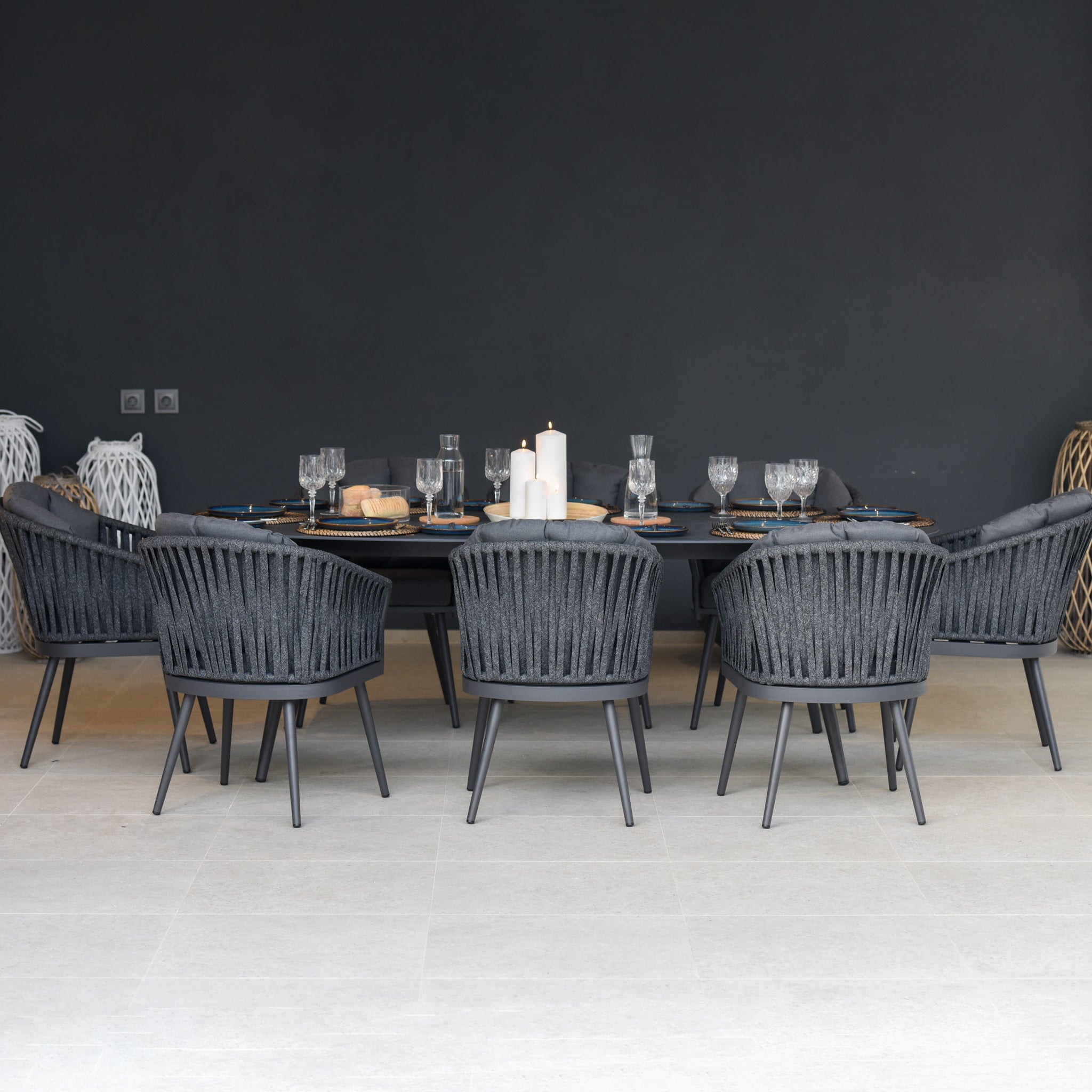 The Monterrey 8 Seat Rope Dining Set with an Oval Ceramic Table in Grey features eight stylish gray woven rope dining chairs arranged around a sleek oval black ceramic table, elegantly set with tableware and candles.