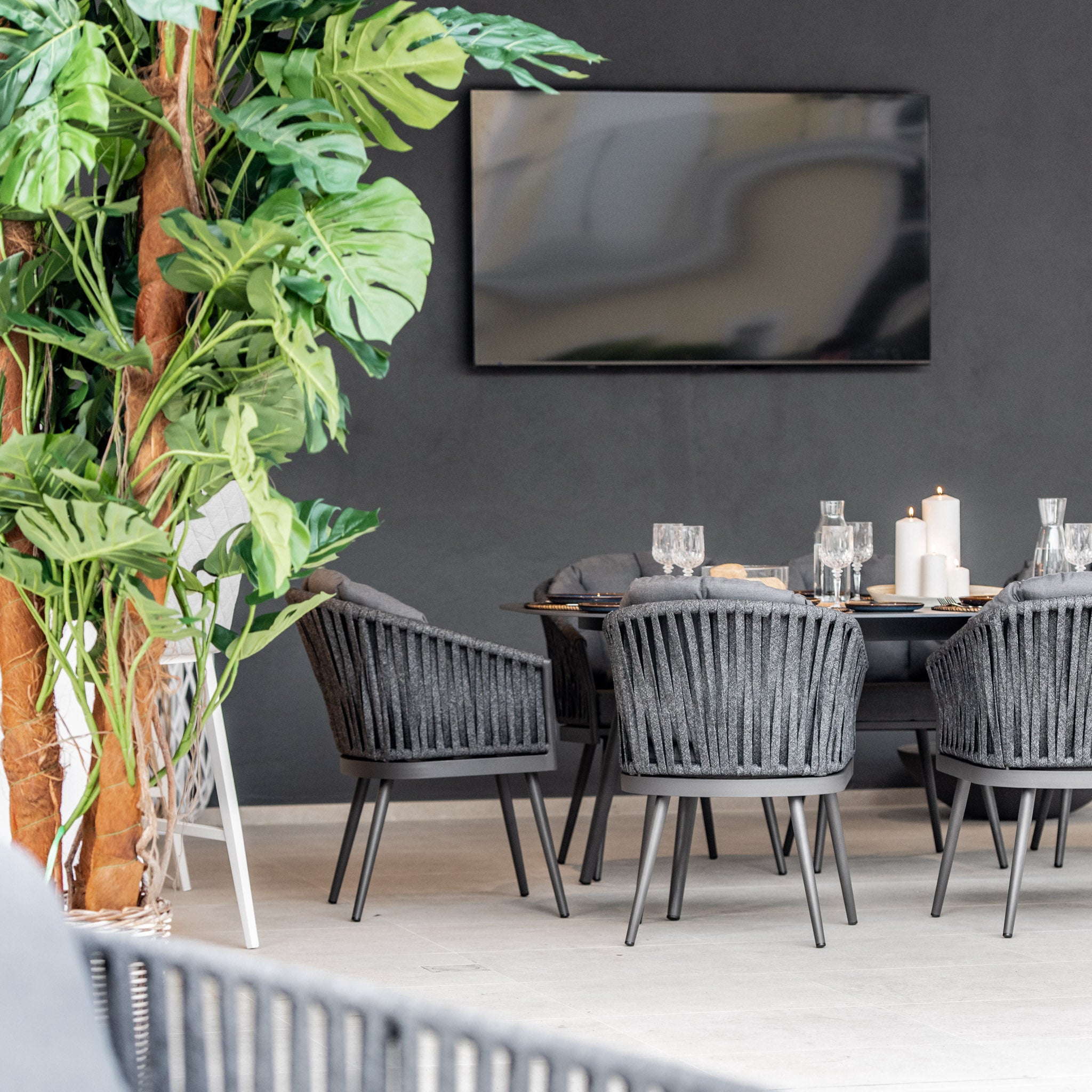 Modern dining area with a Harbour Lifestyle Monterrey 8 Seat Rope Dining Set with an Oval Ceramic Table in Grey, a wall-mounted TV, candles, glassware, and large green plants.
