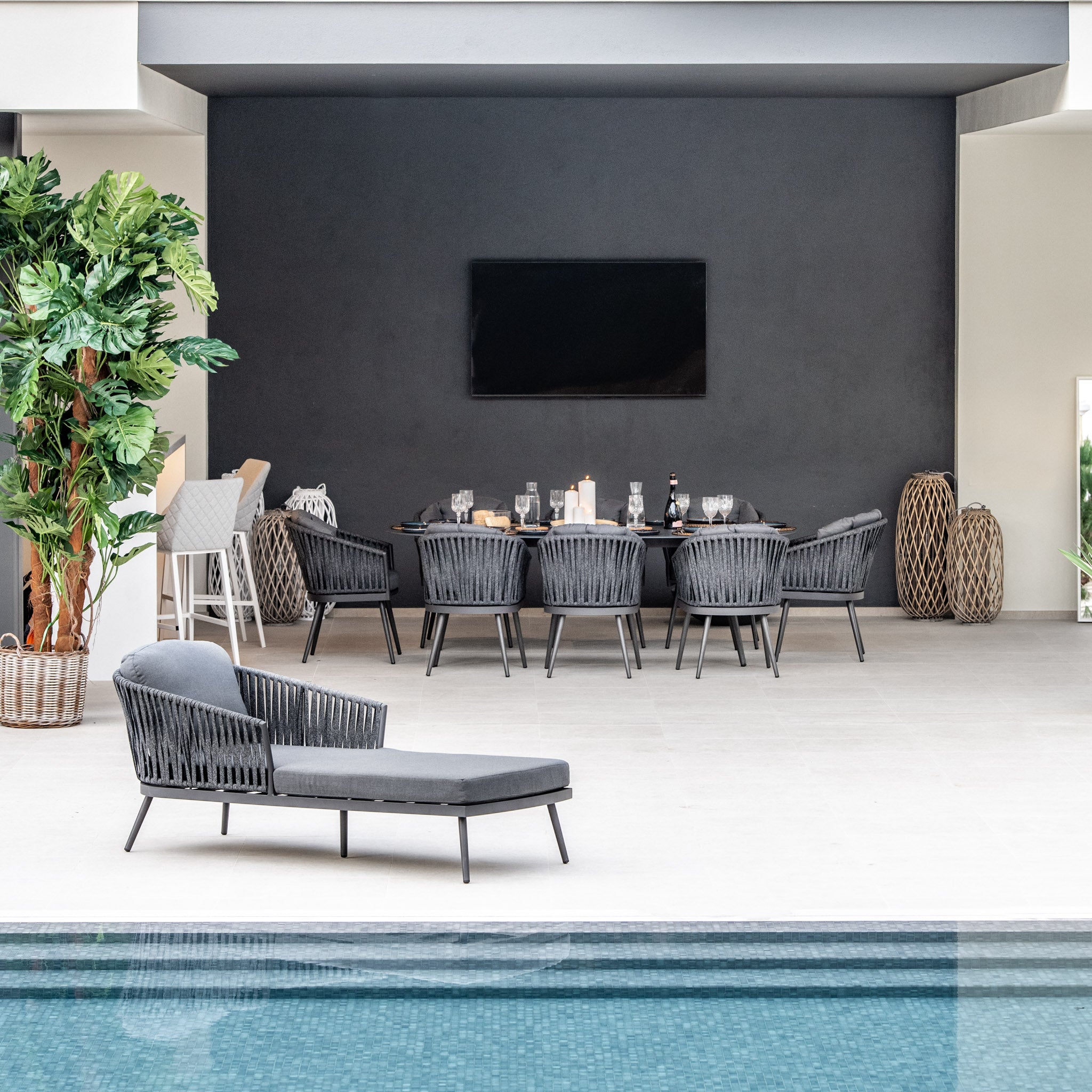 Modern outdoor dining area with a Harbour Lifestyle Monterrey 8 Seat Rope Dining Set with Oval Ceramic Table in Grey, wicker and outdoor rope dining chairs, a chaise lounge by the pool, and a large plant on the left.