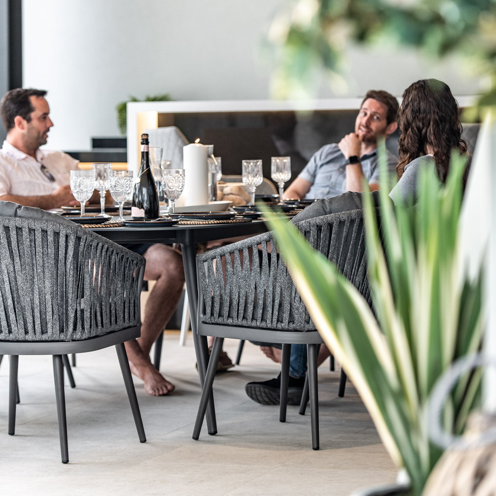 A group of people seated around a Monterrey 8 Seat Rope Dining Set with Oval Ceramic Table in Grey, engaging in lively conversation. The outdoor rope dining chairs provide both comfort and style as friends share stories and laughter over wine and fine glassware.