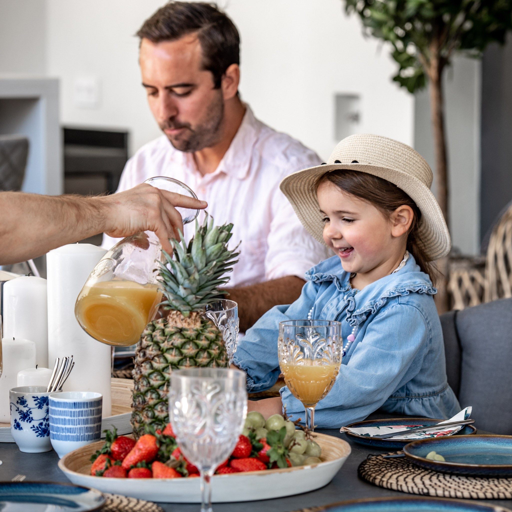 A man and a young girl smiling, sitting at the Harbour Lifestyle Monterrey 8 Seat Rope Dining Set with Oval Ceramic Table in Grey, with fruit and juice being served. The girl is wearing a straw hat, enjoying the Monterrey vibe.