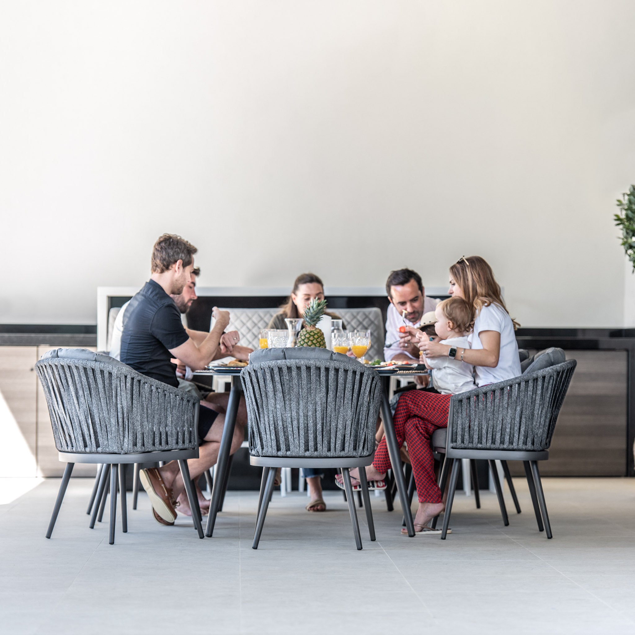 A group of six people are dining at a modern Monterrey 8 Seat Rope Dining Set with an Oval Ceramic Table in Grey, enjoying food and drinks, seated on comfortable outdoor rope dining chairs, engaged in conversation and eating.