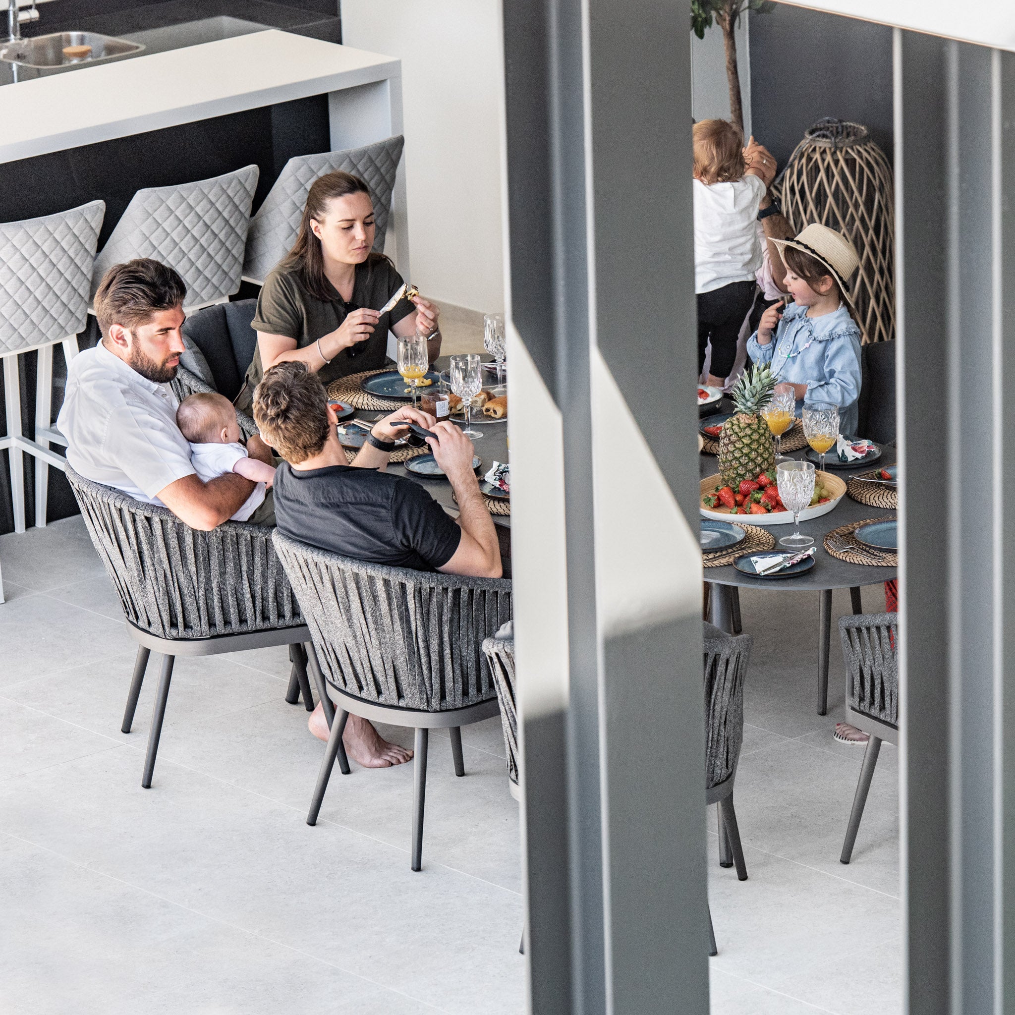 A family of four enjoys a meal together at a Monterrey 8 Seat Rope Dining Set with Oval Ceramic Table in Grey, while another family member stands nearby holding a child, all surrounded by comfortable outdoor rope dining chairs.