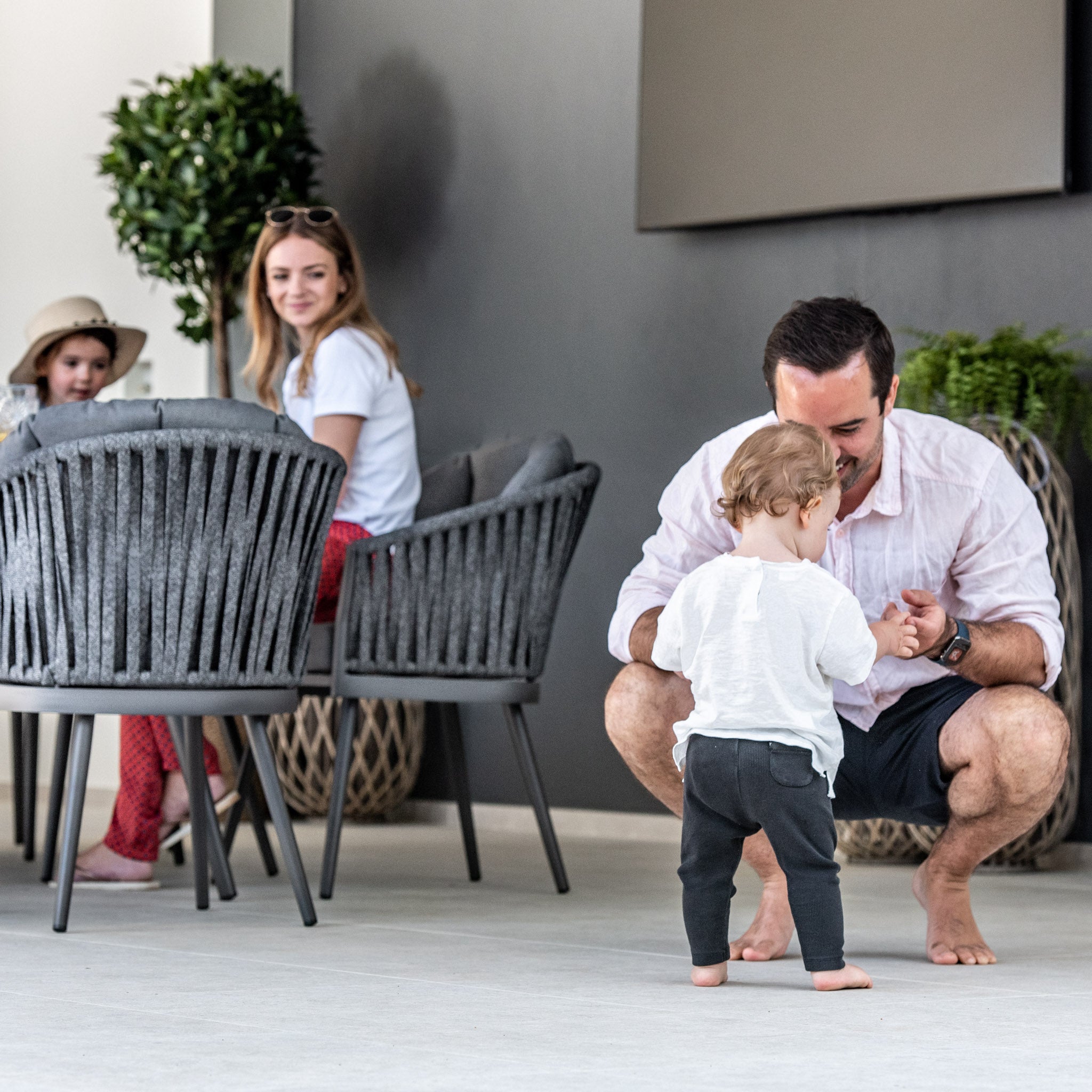 A toddler taking first steps, encouraged by a man, with two women seated at the Monterrey 8 Seat Rope Dining Set with Oval Ceramic Table in Grey. In the background, the outdoor rope dining chairs from Harbour Lifestyle Monterrey add a touch of elegance to the scene.