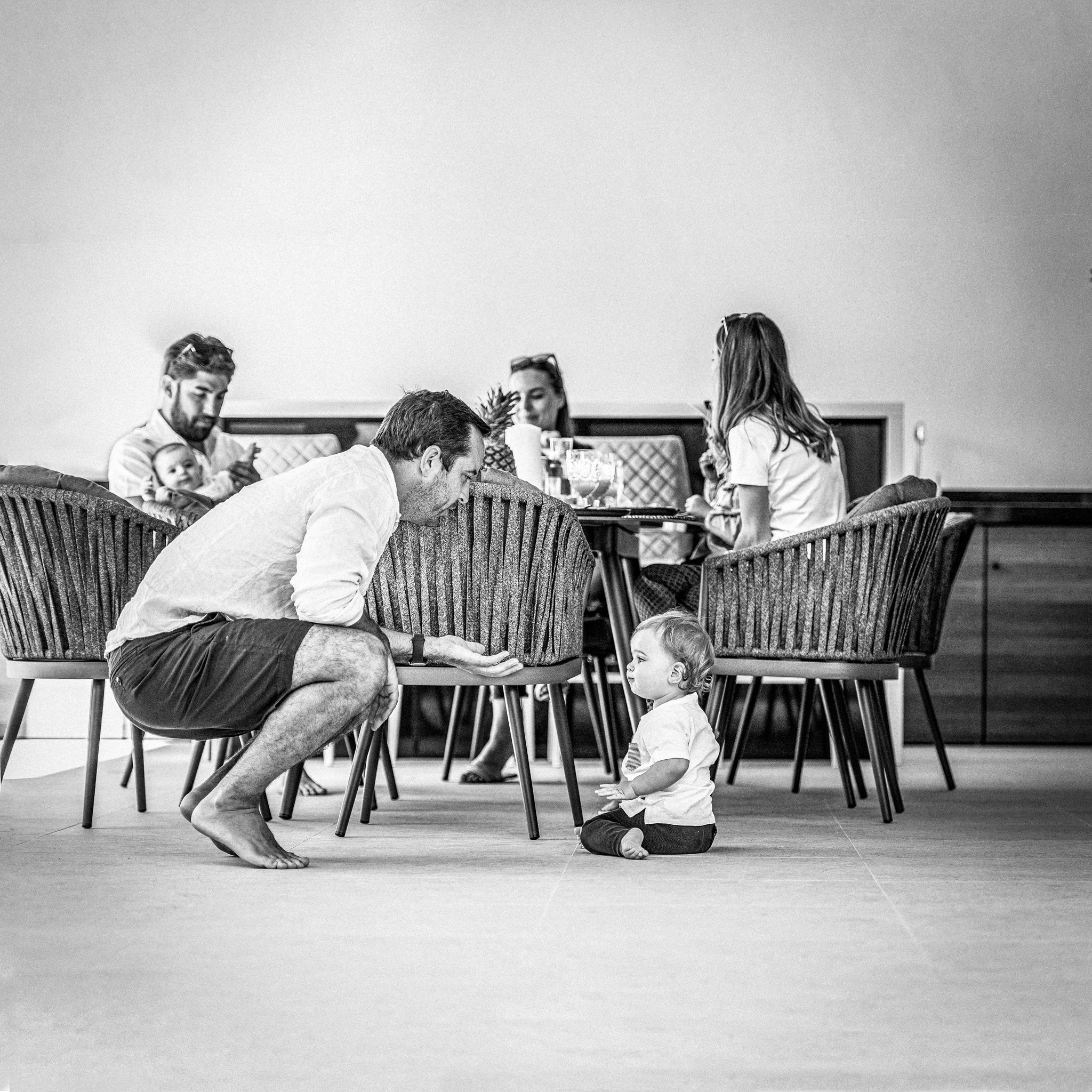 A man crouches to interact with a baby sitting on the floor, while others are seated at the Monterrey 8 Seat Rope Dining Set with Oval Ceramic Table in Grey in the background. Black and white photo.