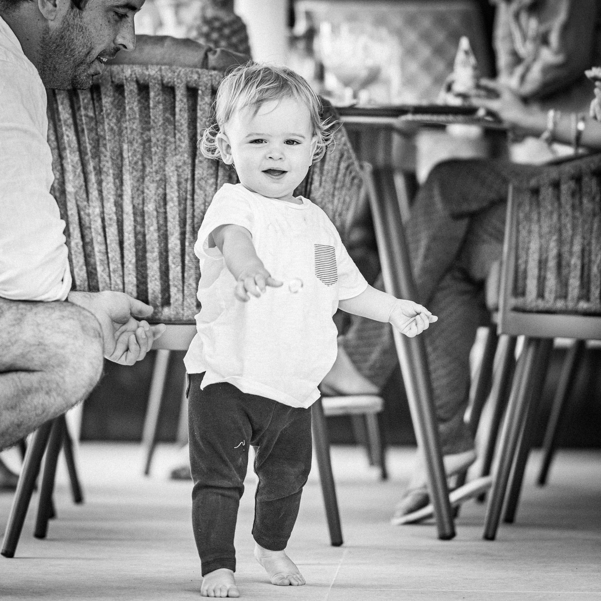A black and white image captures a toddler walking barefoot indoors, with an adult crouching nearby and seated people in the background. The scene exudes the timeless elegance typical of Harbour Lifestyle settings, reminiscent of afternoons around the Monterrey 8 Seat Rope Dining Set with Oval Ceramic Table in Grey.
