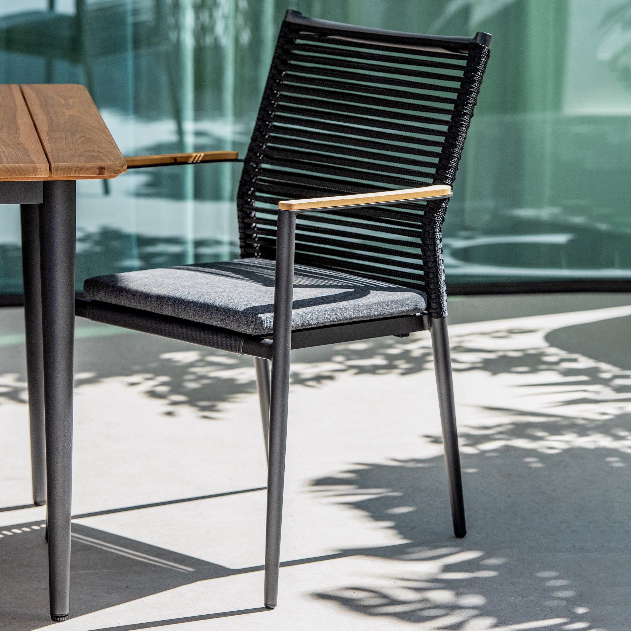A modern black metal chair with a cushioned seat beside a teak table outdoors, casting shadows on the ground. The setup pairs perfectly with the Portland 6 Seat Rectangular Dining Set with Teak Table in Charcoal, featuring weatherproof aluminium legs for durability.