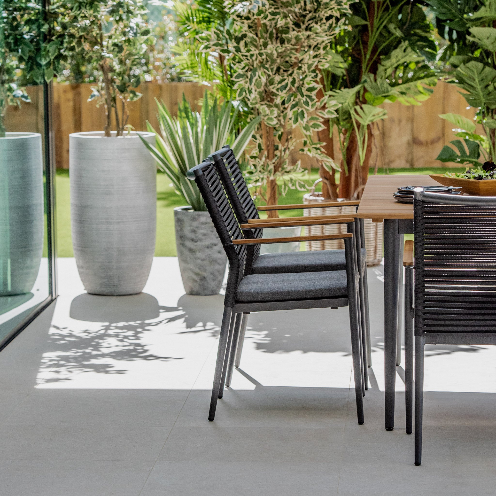 Modern outdoor patio featuring the Portland 6 Seat Rectangular Dining Set with Teak Table in Charcoal, black chairs with weatherproof aluminium legs, and lush green plants in large gray planters.
