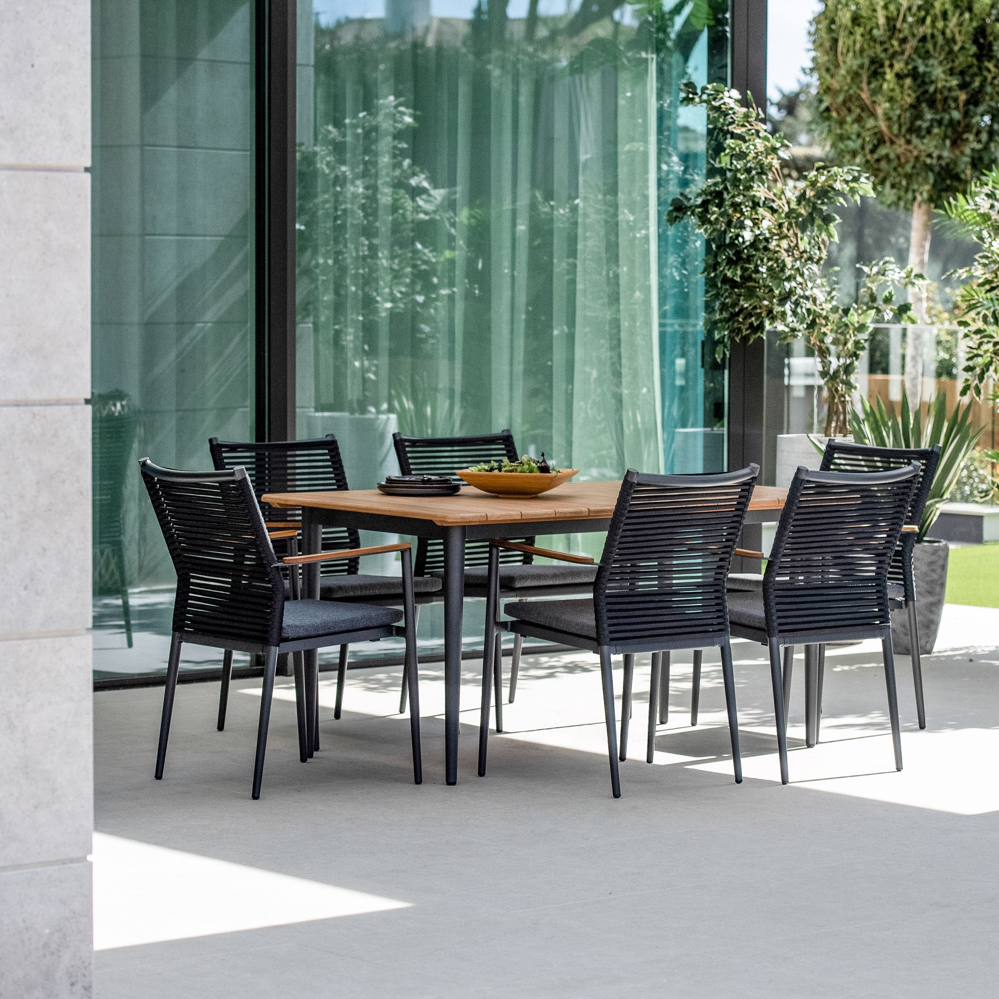 Outdoor dining area featuring the Portland 6 Seat Rectangular Dining Set with a teak table in charcoal and black chairs, set against a modern glass wall backdrop, all supported by weatherproof aluminium legs and surrounded by lush greenery.