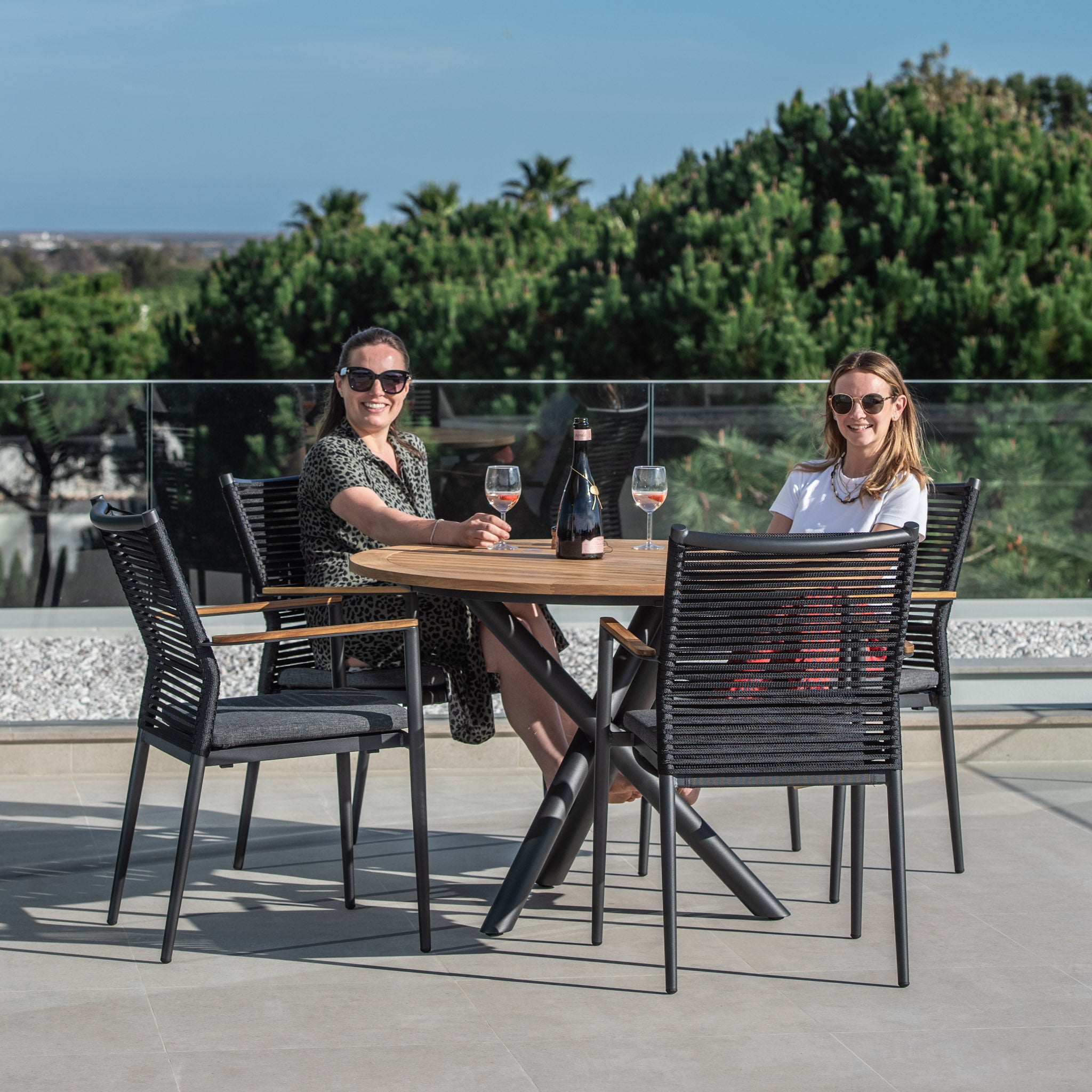 Two women sit at a Portland 4 Seat Round Dining Set with Teak Table in Charcoal, with wine glasses in hand, enjoying a sunny day. The scene features a scenic view in the background, and they're comfortably seated on flat rope weaved armchairs around the stylish teak wood table.