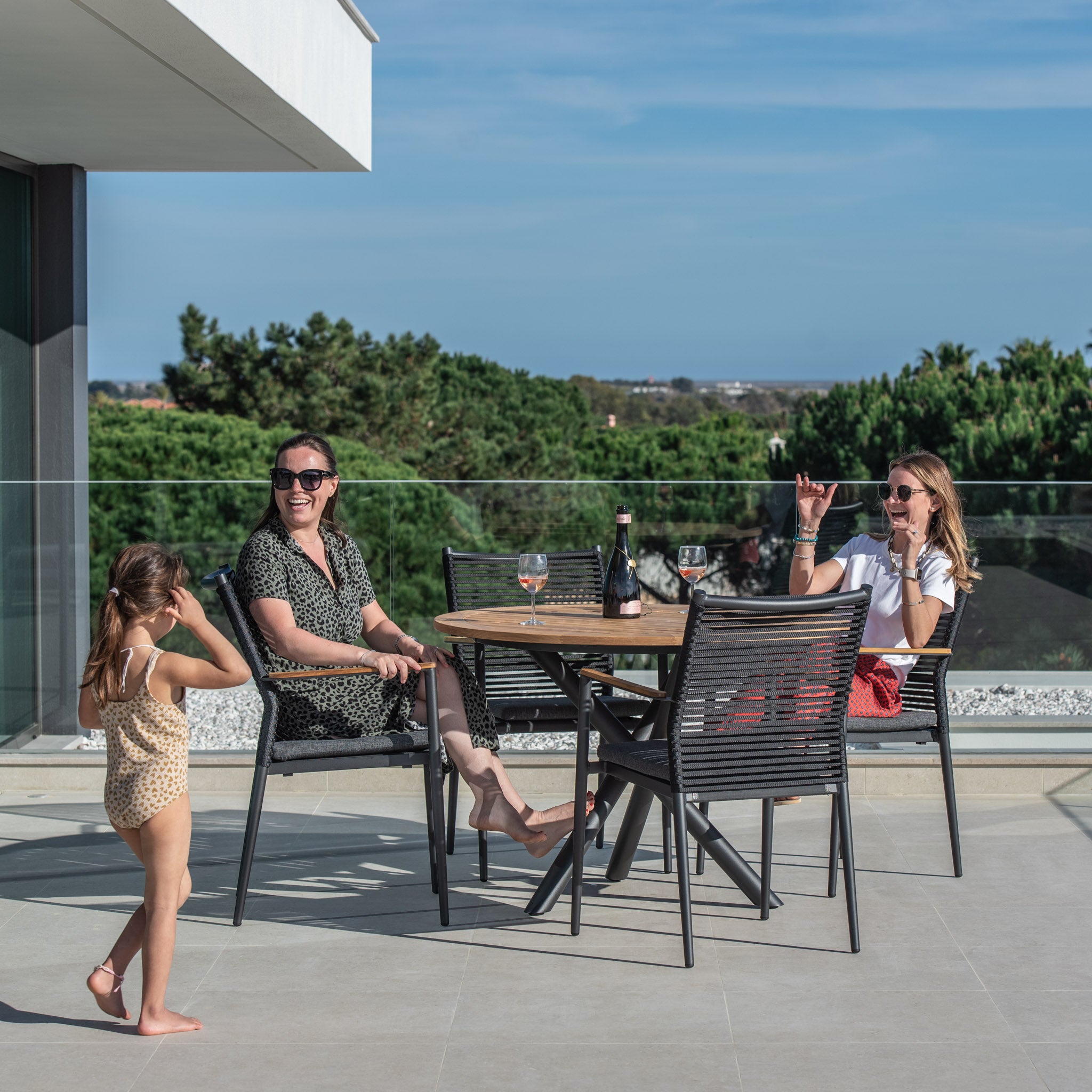 Two women sit at the Portland 4 Seat Round Dining Set with Teak Table in Charcoal, drinks in hand, smiling, while a child in swimwear stands nearby on a sunny day. They enjoy the comfort of flat rope weaved armchairs as they share laughs and create memories by their outdoor dining set.