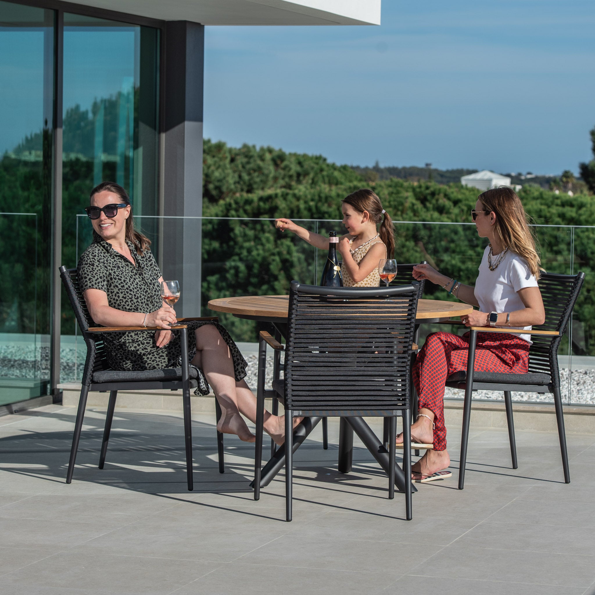 Three women sitting at the Portland 4 Seat Round Dining Set with Teak Table in Charcoal, talking and laughing on a sunny day, with trees and buildings in the background. They are comfortably seated in flat rope weaved armchairs around the beautiful teak wood table.