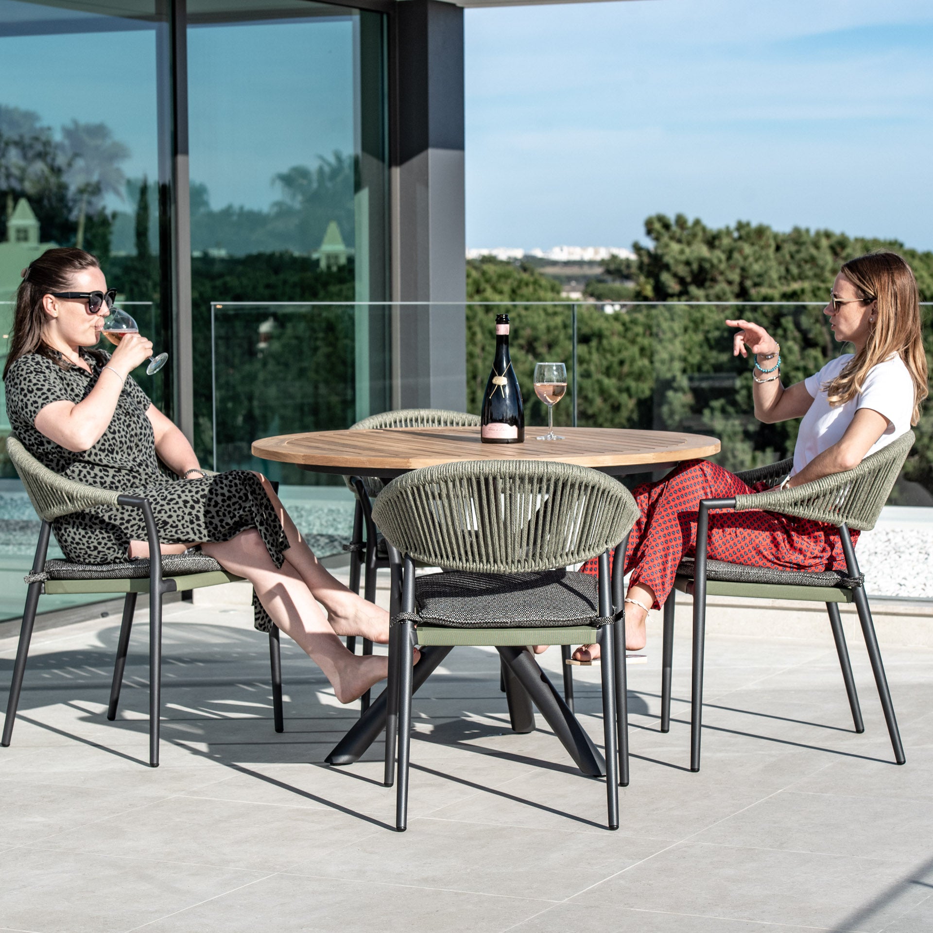 Two women sit at the Cloverly 4 Seat Round Dining Set with a Teak Table Top in Olive Green, enjoying drinks and a bottle of wine while soaking up the sun on a balcony adorned with stylish woven chairs.