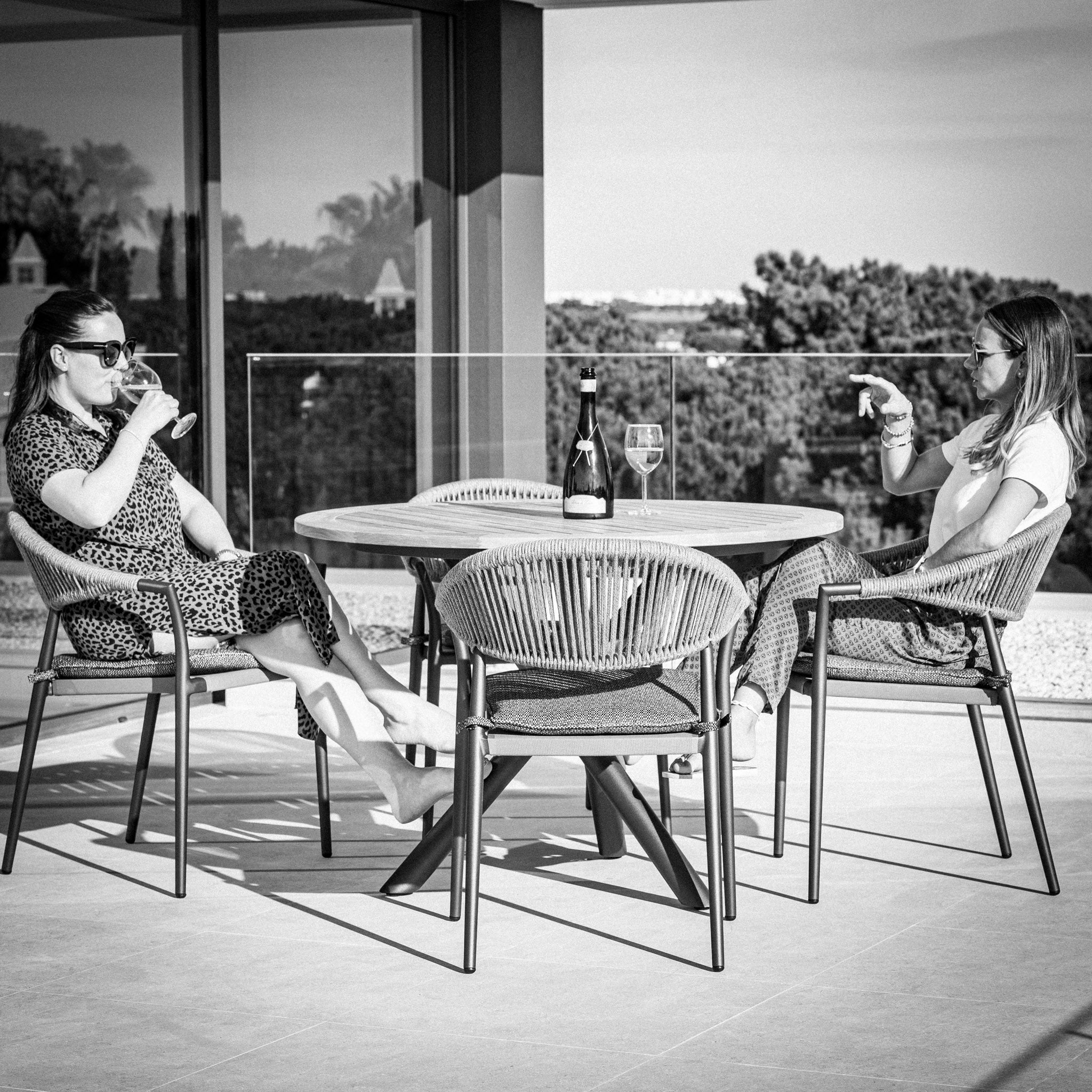 Two women sitting at the Cloverly 4 Seat Round Dining Set with a Teak Table Top in Charcoal, enjoying drinks and a conversation on a sunny patio.