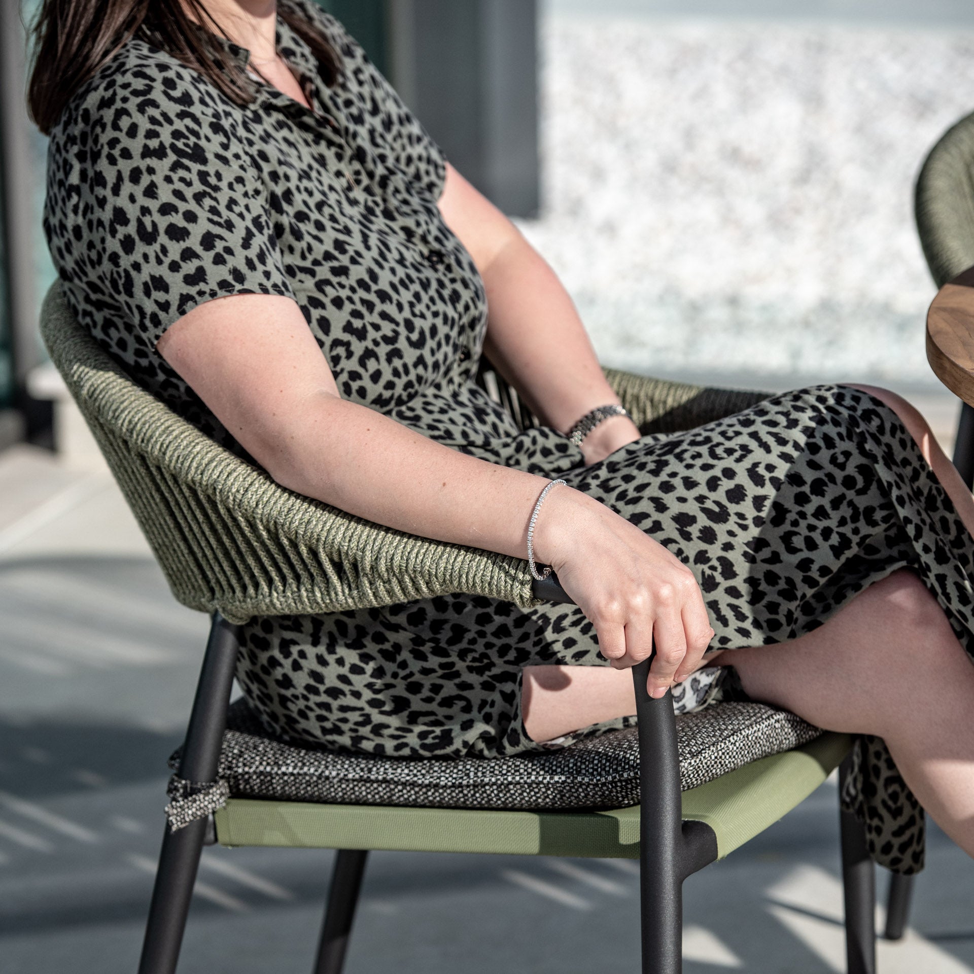 Person in a leopard print dress sitting on a green woven chair outdoors, with hands resting on the armrests beside the Cloverly 4 Seat Round Dining Set with Teak Table Top in Olive Green.