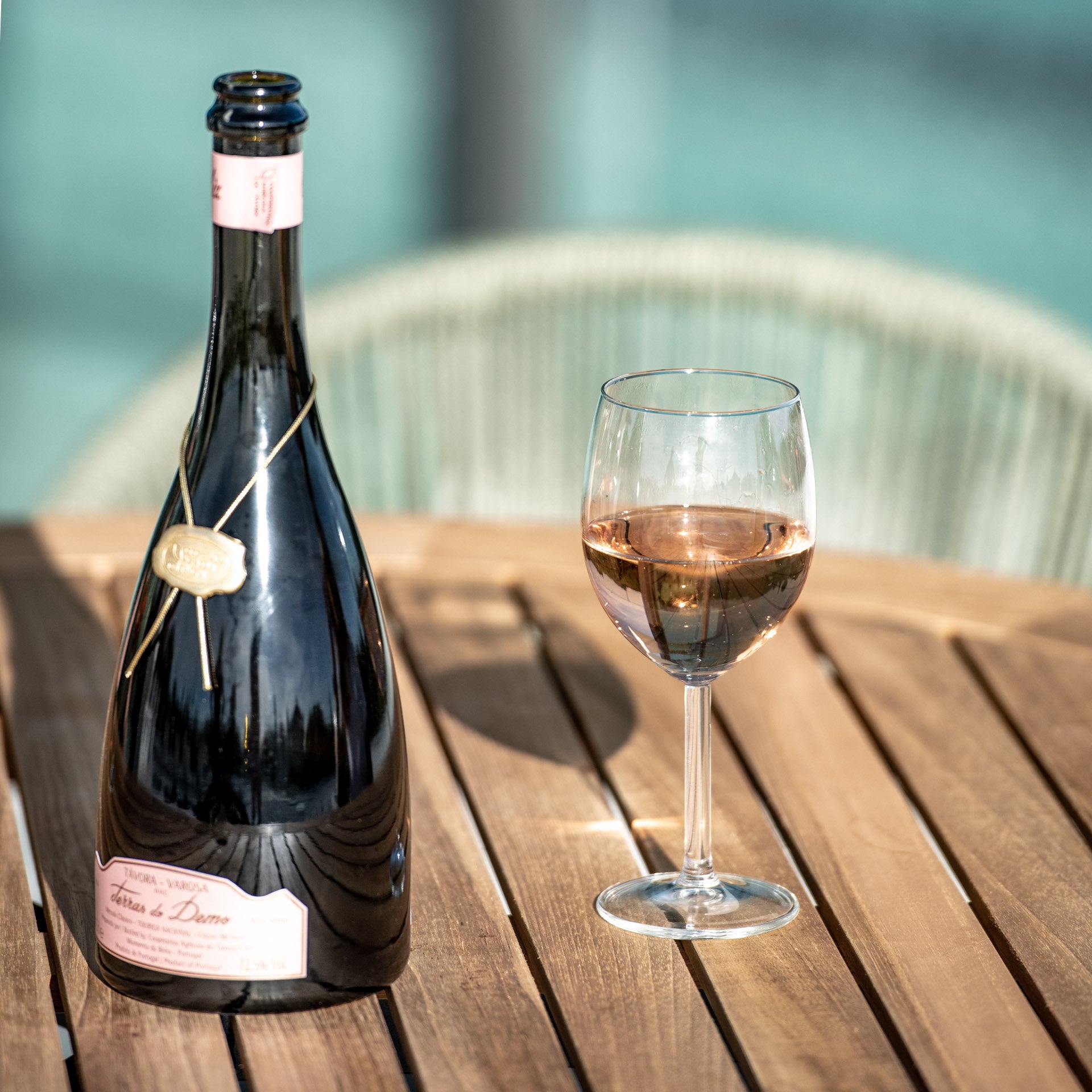 A bottle of rosé wine and a filled wine glass sit gracefully on the teak table top of the Cloverly 4 Seat Round Dining Set in Olive Green, surrounded by woven chairs in an inviting outdoor setting.