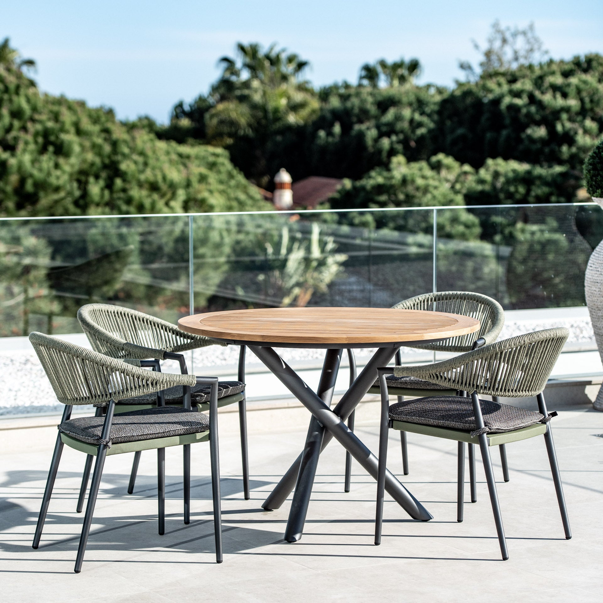 The Cloverly 4 Seat Round Dining Set, featuring a teak table top in Olive Green and four weather-proof aluminum woven chairs, overlooks a lush landscape with trees and clear skies.