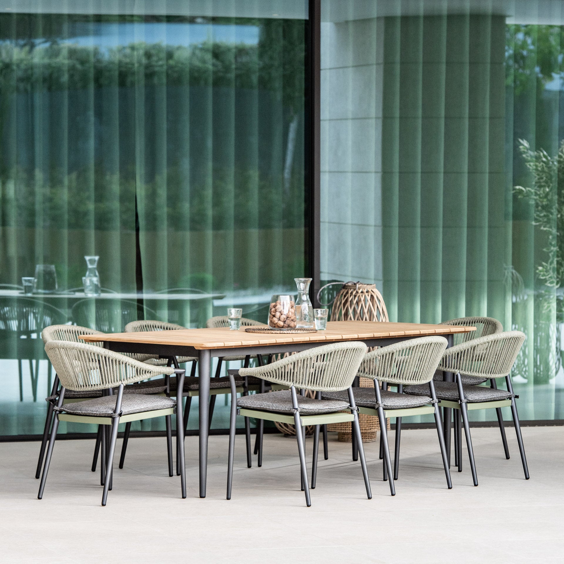 The Cloverly 8 Seat Rectangular Dining with Teak Table in Olive Green features a teak top and black legs, accompanied by six rope-woven chairs for a modern patio look. This dining set is enhanced with an aluminium frame for added durability and style.