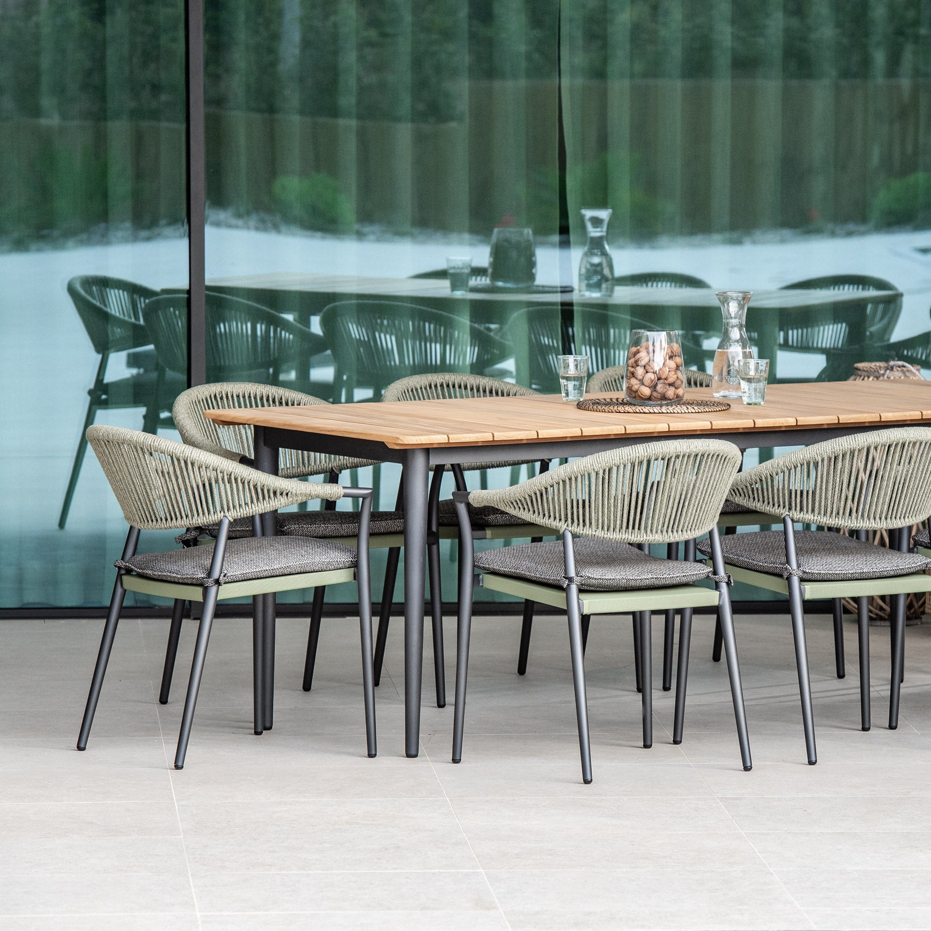The elegant Cloverly 8 Seat Rectangular Dining Set with a teak top table in olive green features aluminum frame construction for durability. This outdoor dining set, complete with coordinating wicker chairs, is situated on a patio in front of a reflective glass wall.