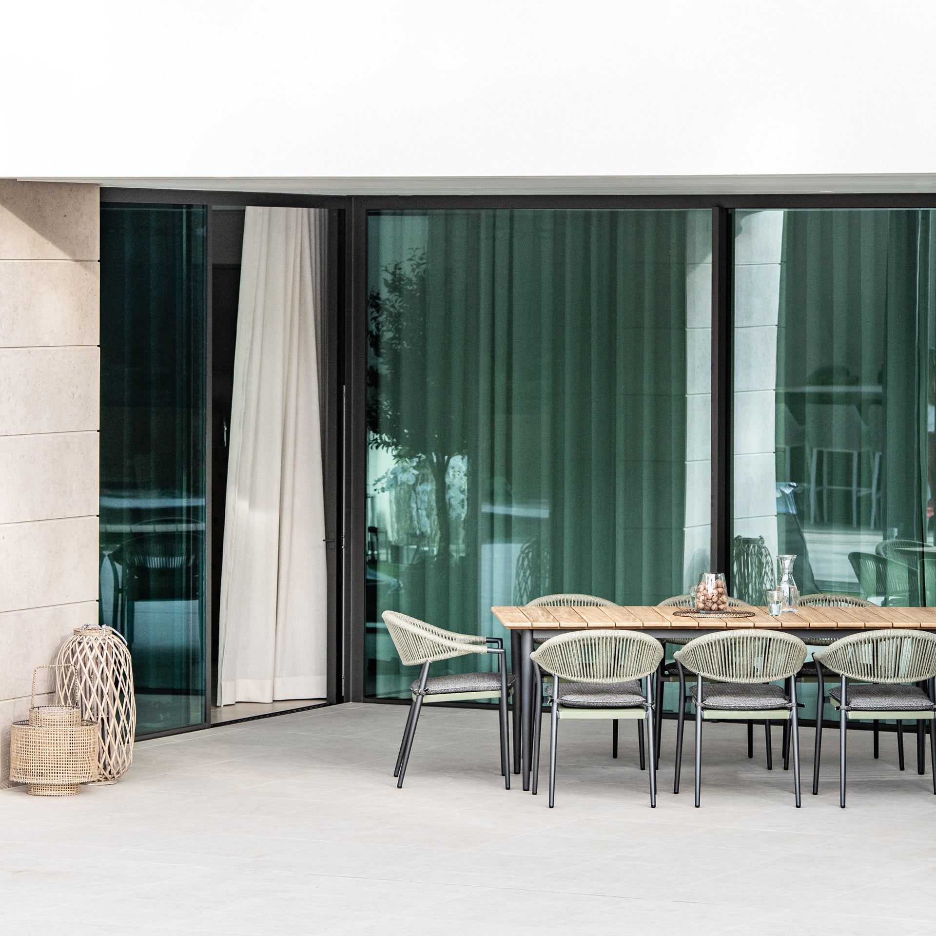 Modern patio featuring the Cloverly 8 Seat Rectangular Dining with Teak Table in Olive Green, complemented by glass doors, green chairs, and wicker decor items beside the door.