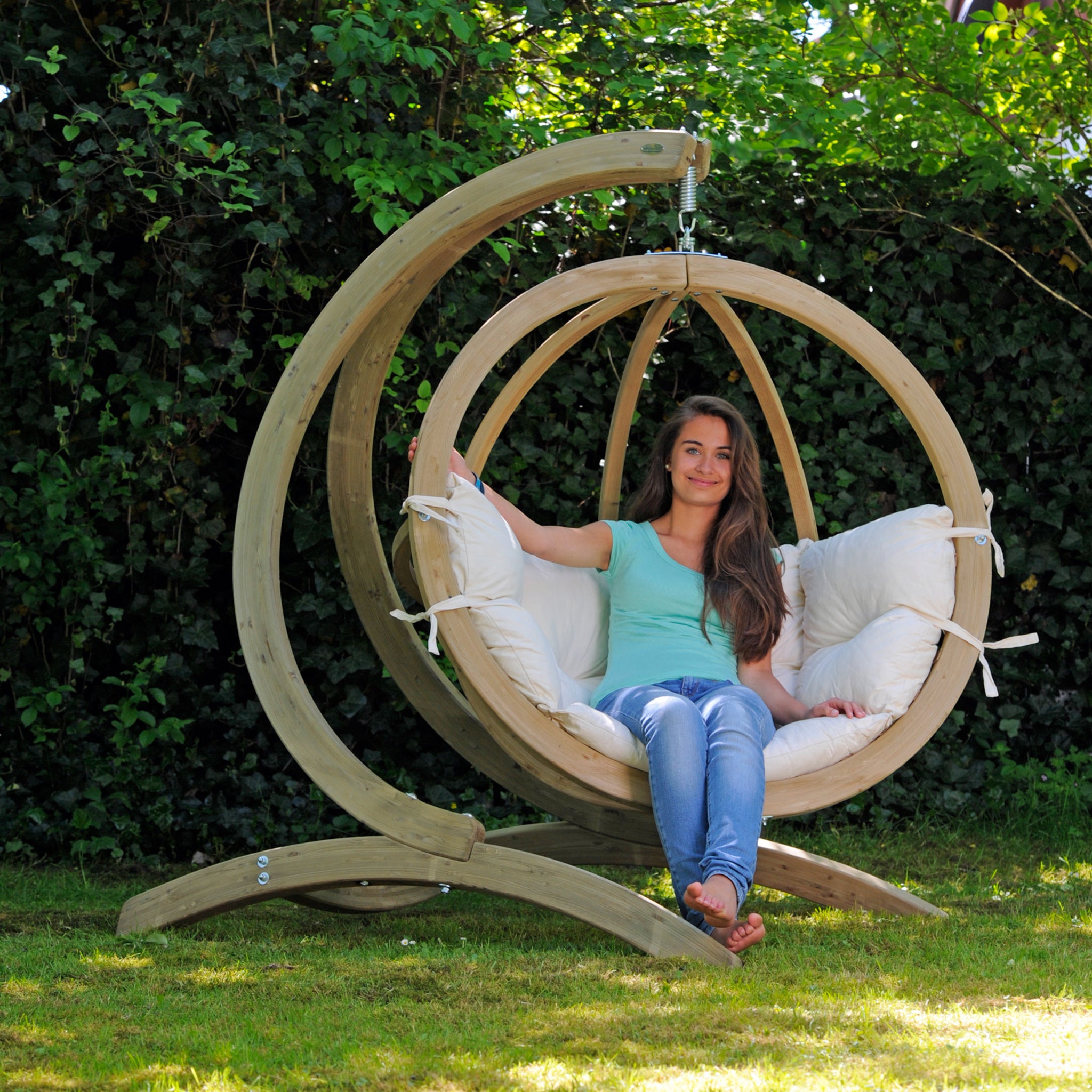 A woman sits in a stylish Natura Globo Single Hanging Chair with a white cushion, set in a garden with lush green foliage in the background. This comfortable chair seamlessly blends indoor and outdoor living spaces.