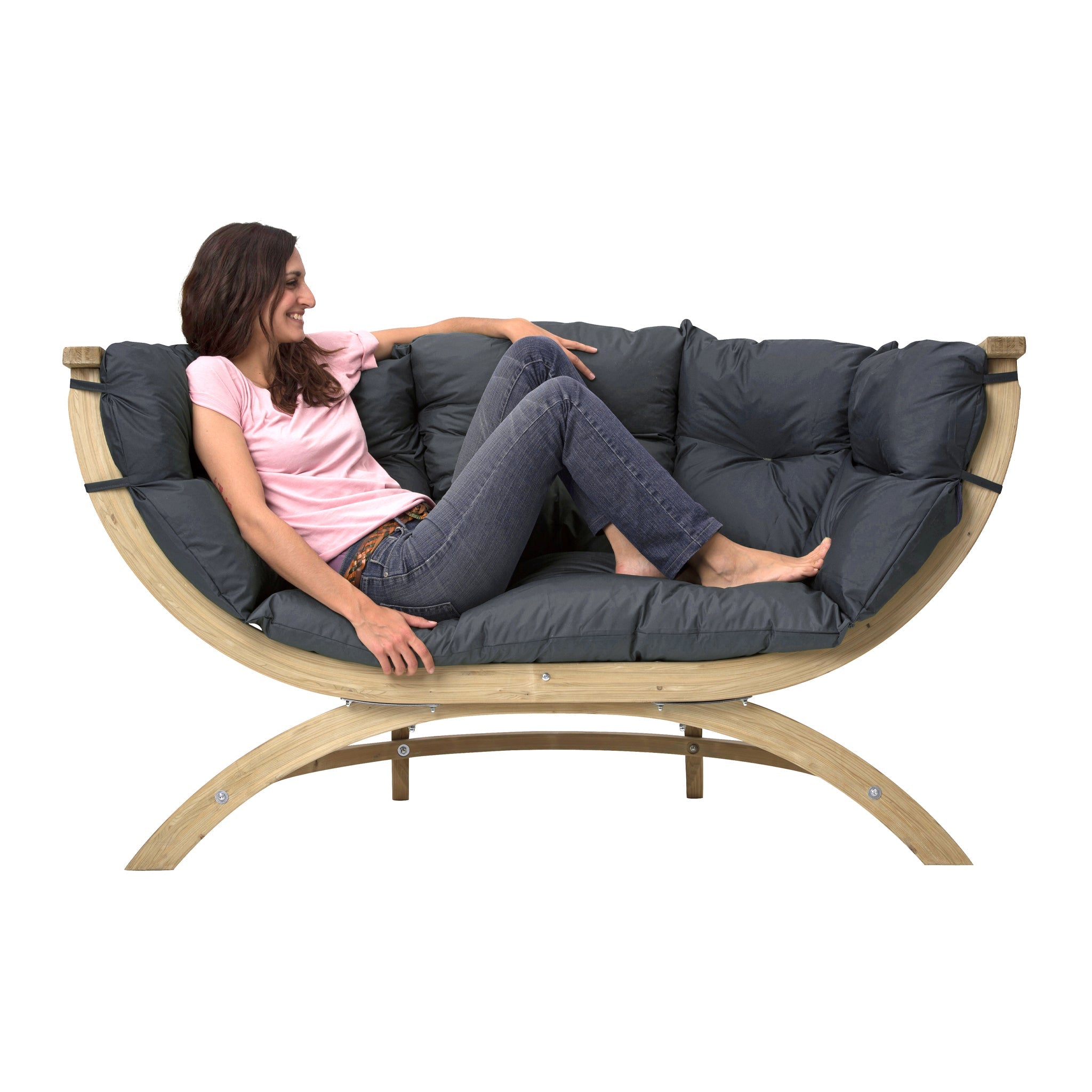 A woman in a pink shirt and jeans is lounging on the Globo Siena Due Seat Weatherproof in Anthracite Grey, a curved wooden bench with upholstered cushions crafted from sustainably sourced wood.