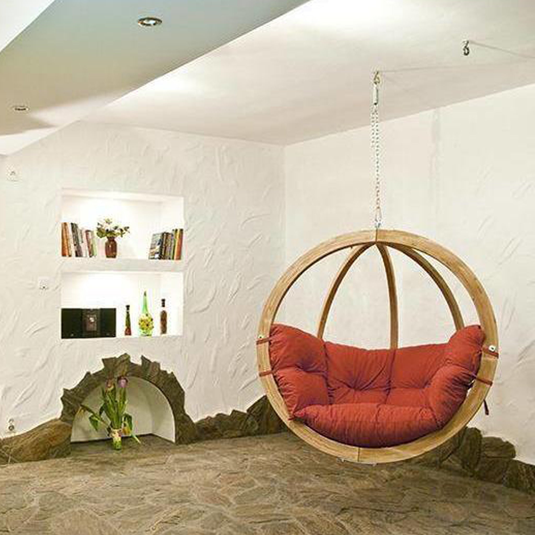 Room with a Natura Globo Single Hanging Chair featuring a red cushion, built-in wall shelves filled with books and decor items, stylish furniture, and stone flooring that seamlessly connects indoor and outdoor living spaces.