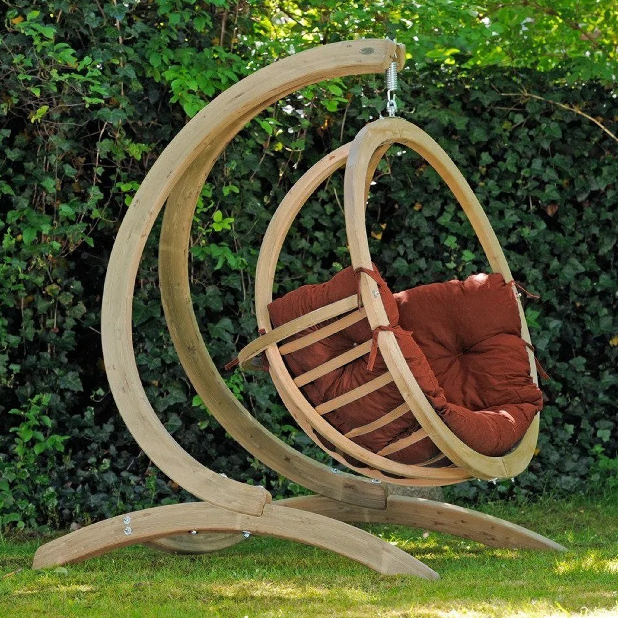 The Natura Globo Single Hanging Chair, complete with comfortable red cushions, is stylishly suspended from a wooden frame in a lush green garden, making it perfect for both indoor and outdoor living spaces.