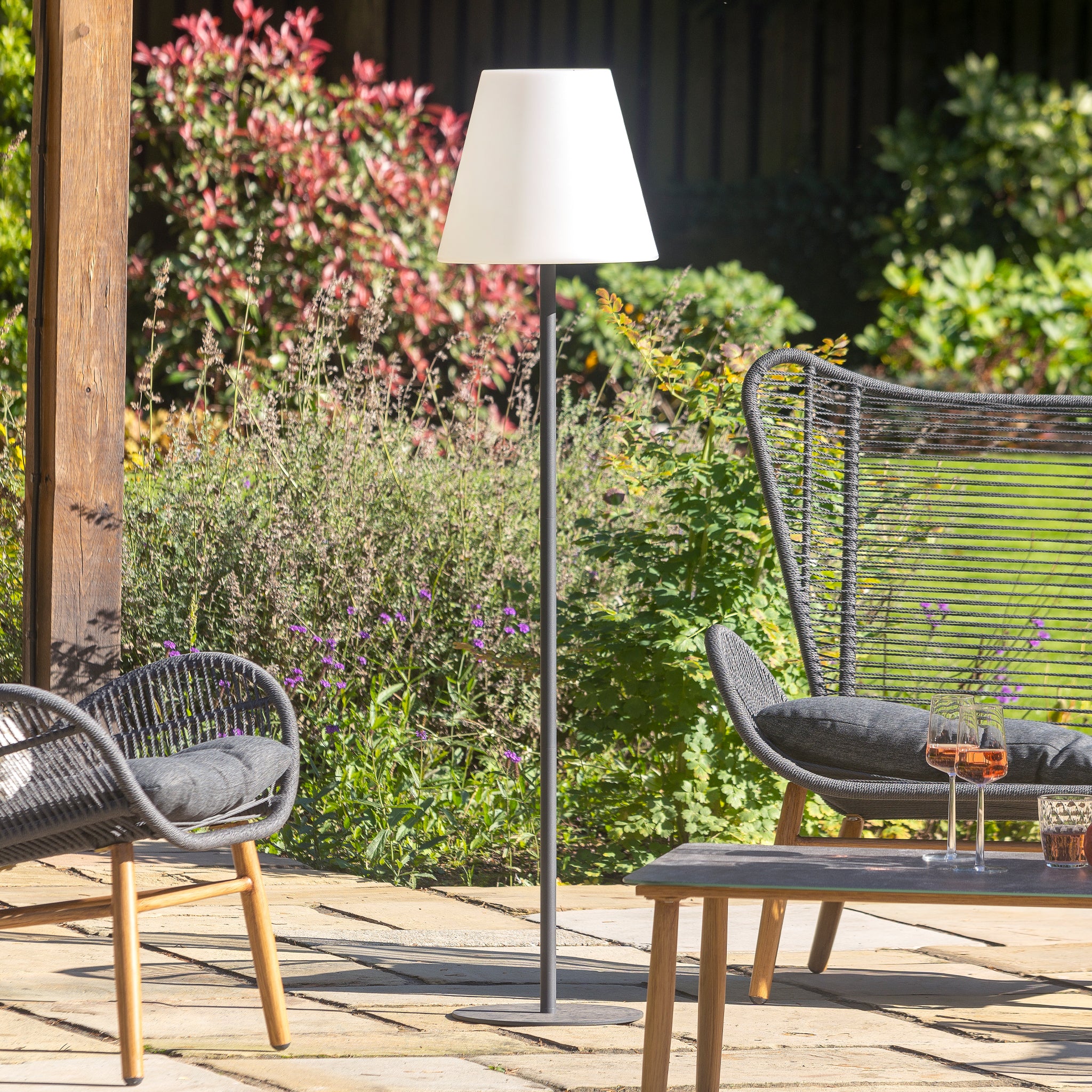 A serene patio with chairs, a table, and the chic Pacific Lifestyle grey metal outdoor and indoor floor lamp. Encircled by greenery under a charming wooden pergola, this setup adds both style and function to your outdoor area.