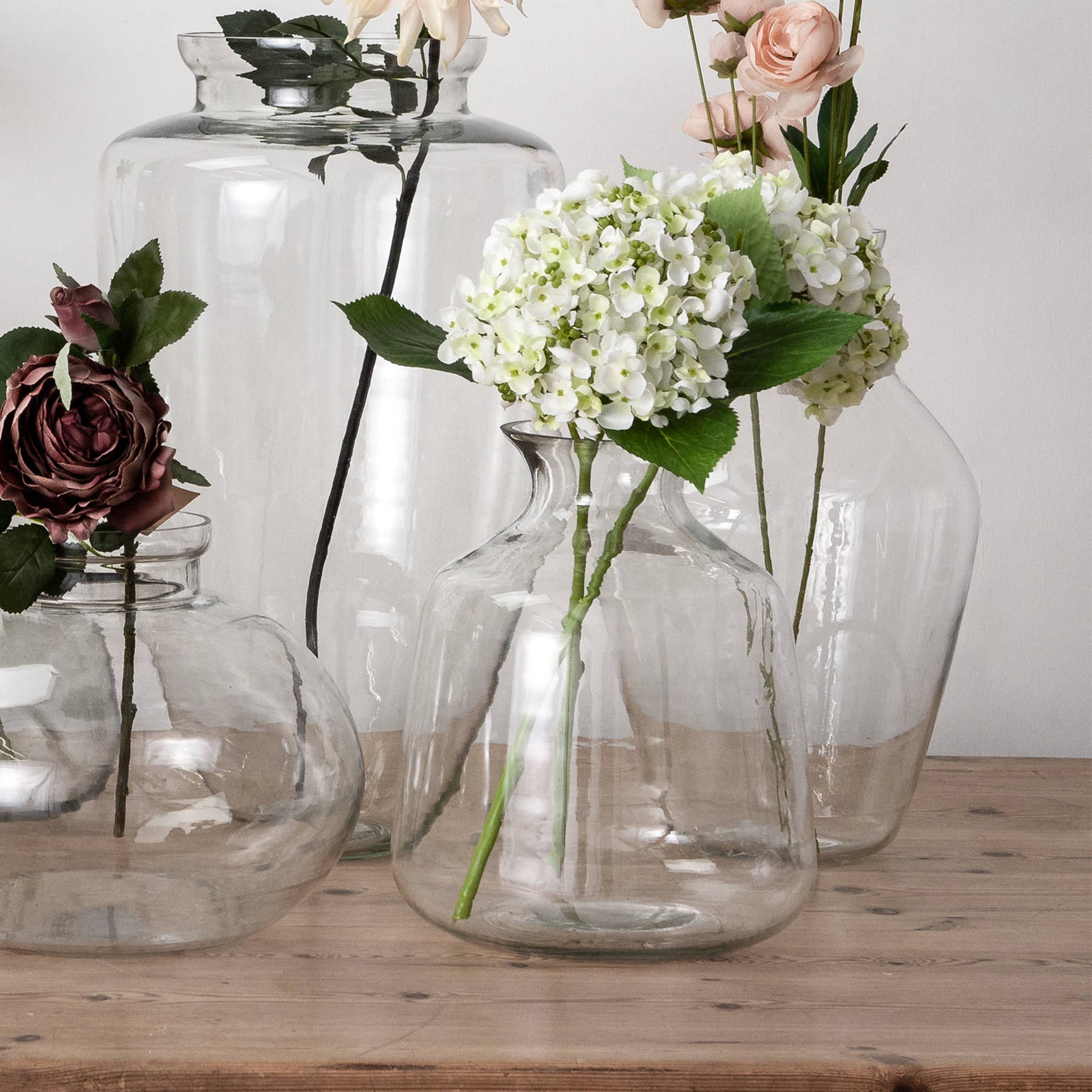 On a wooden surface lie decorative glass vases of various shapes and sizes, each filled with colorful flowers such as roses and hydrangeas. Standing out among them is the striking Hydria Glass Vase, which introduces an ancient Greek touch to the elegant display.