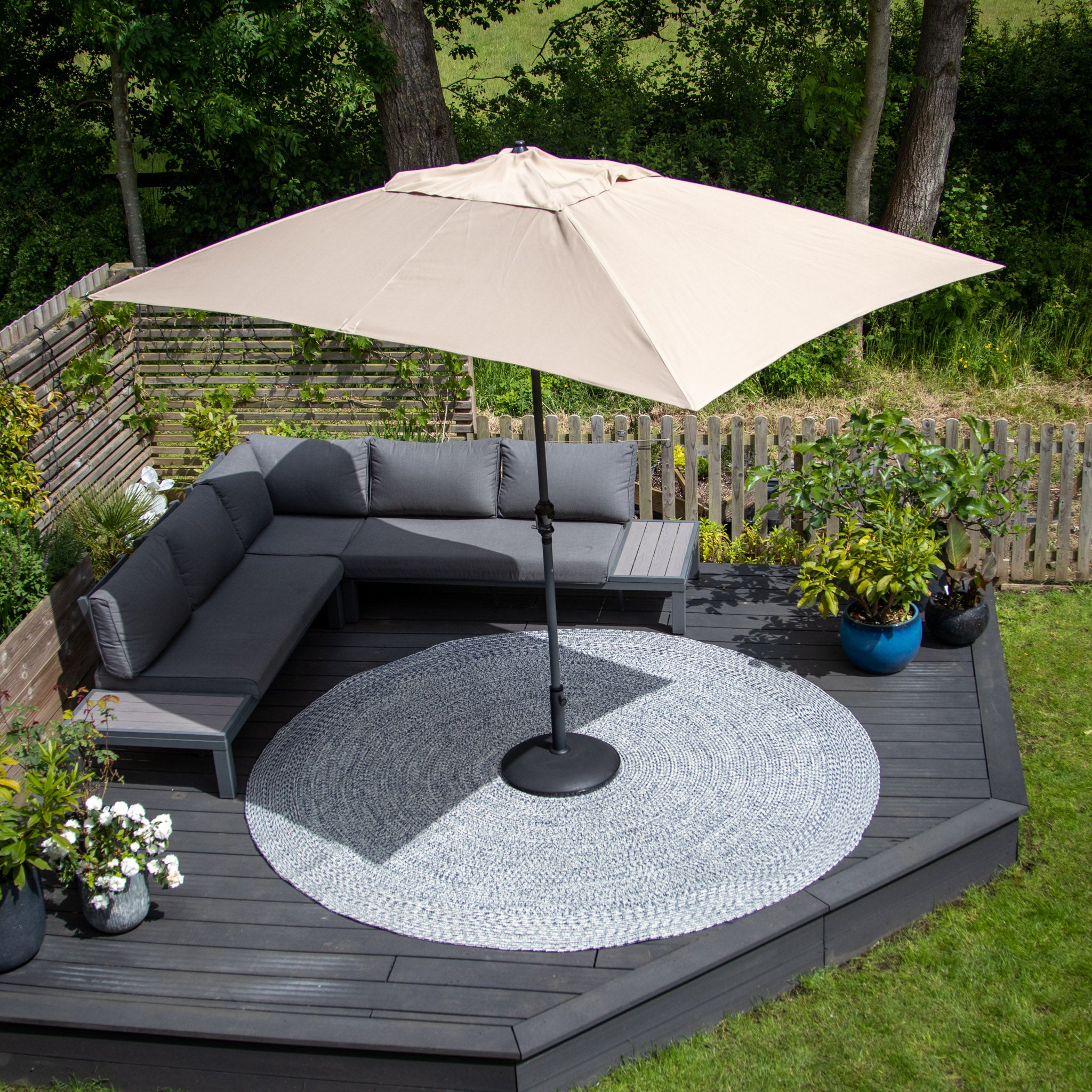 A cozy outdoor patio with a large beige umbrella, sectional sofa, and an Apollo 300cm Round Indoor and Outdoor Rug in Prussian Blue, surrounded by greenery and flowers. This weatherproof, UV-resistant rug perfectly complements the inviting atmosphere.