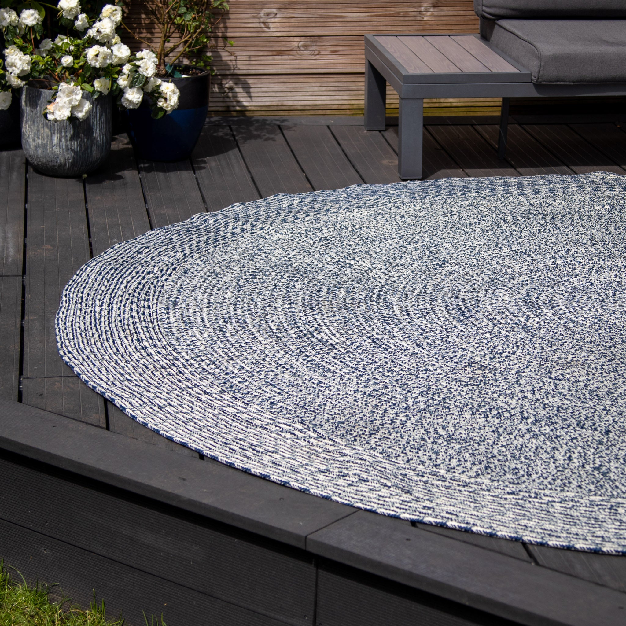 The Apollo 300cm Round Indoor and Outdoor Rug in Prussian Blue adds a touch of elegance to the dark wooden deck, featuring a round, woven design beside a potted plant and grey patio furniture. This weatherproof, UV-resistant rug is as functional as it is stylish.