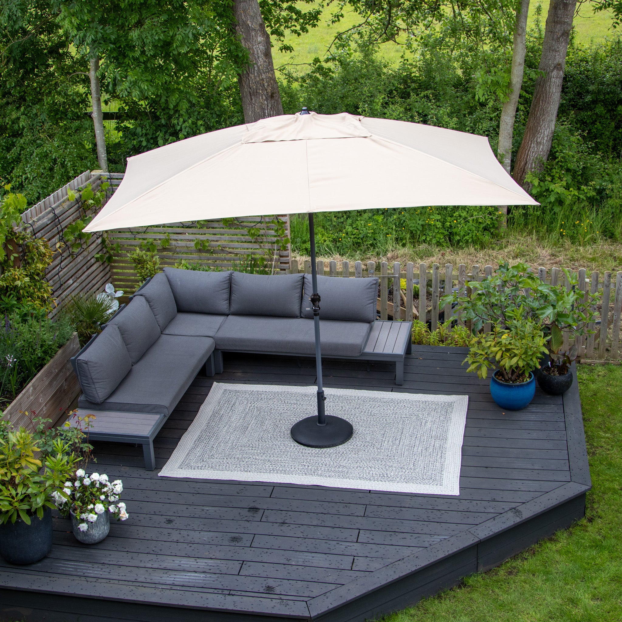 A backyard deck featuring a gray sectional sofa under a large beige umbrella, complemented by the Hades 290cm x 190cm Weatherproof Indoor and Outdoor Rug in Cobble Grey, and surrounded by lush plants. This serene spot is not only stylish but also UV resistant, ensuring long-lasting beauty.
