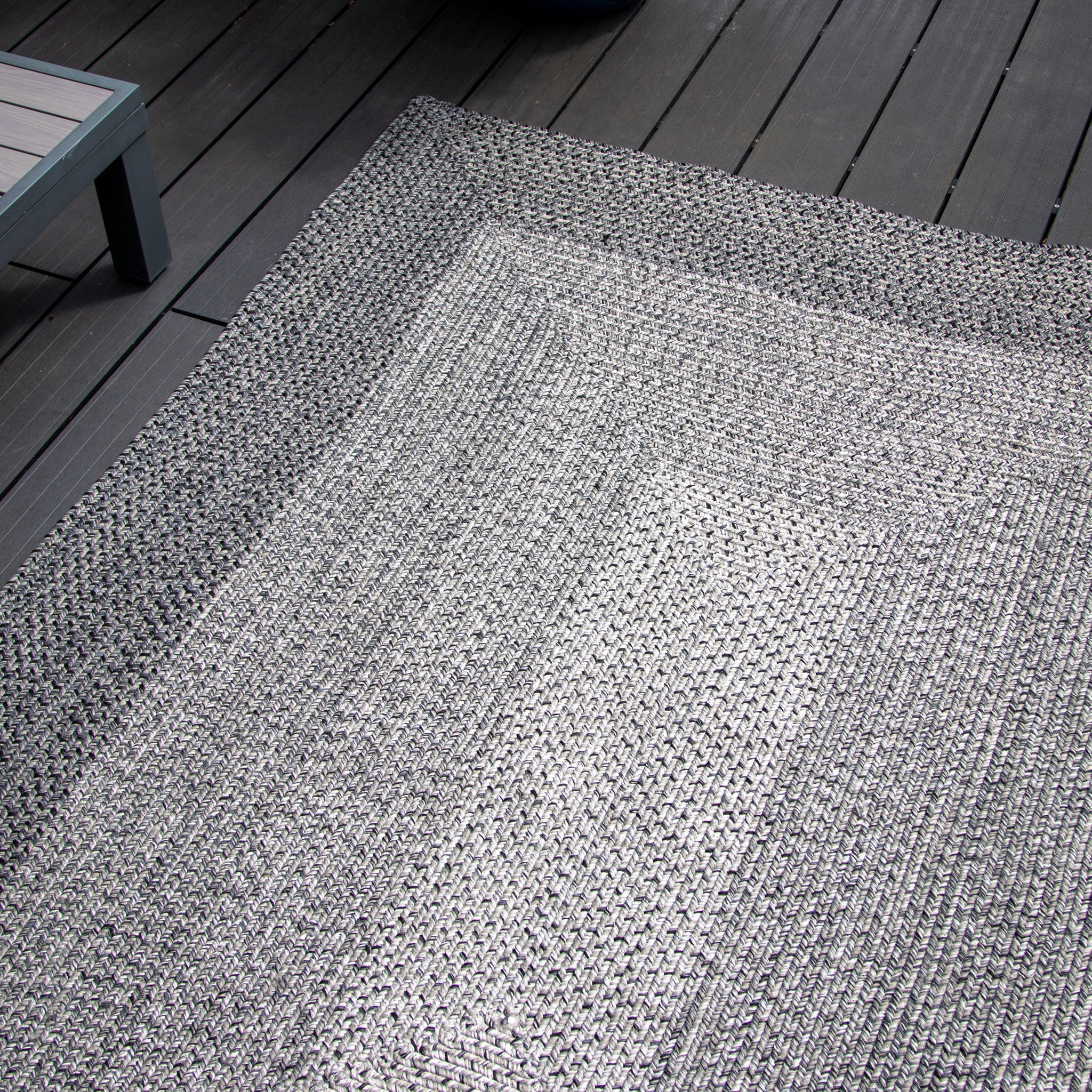 An Artemis 366cm x 274cm Weatherproof Indoor and Outdoor Rug in Slate Grey on a dark gray wooden deck near a blue and gray weatherproof bench.