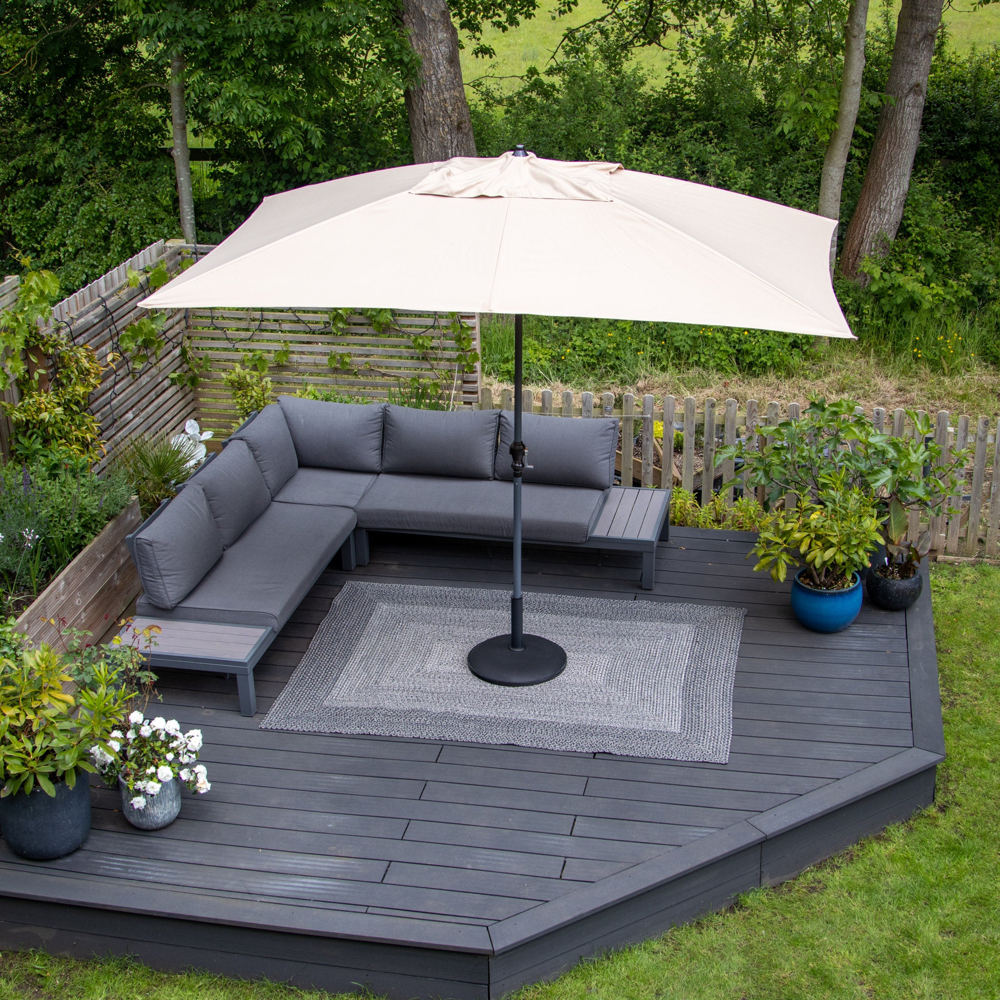 A patio with a gray sectional sofa, a large beige umbrella, weatherproof plants, and the Artemis 366cm x 274cm Weatherproof Indoor and Outdoor Rug in Slate Grey on a wooden deck surrounded by trees.