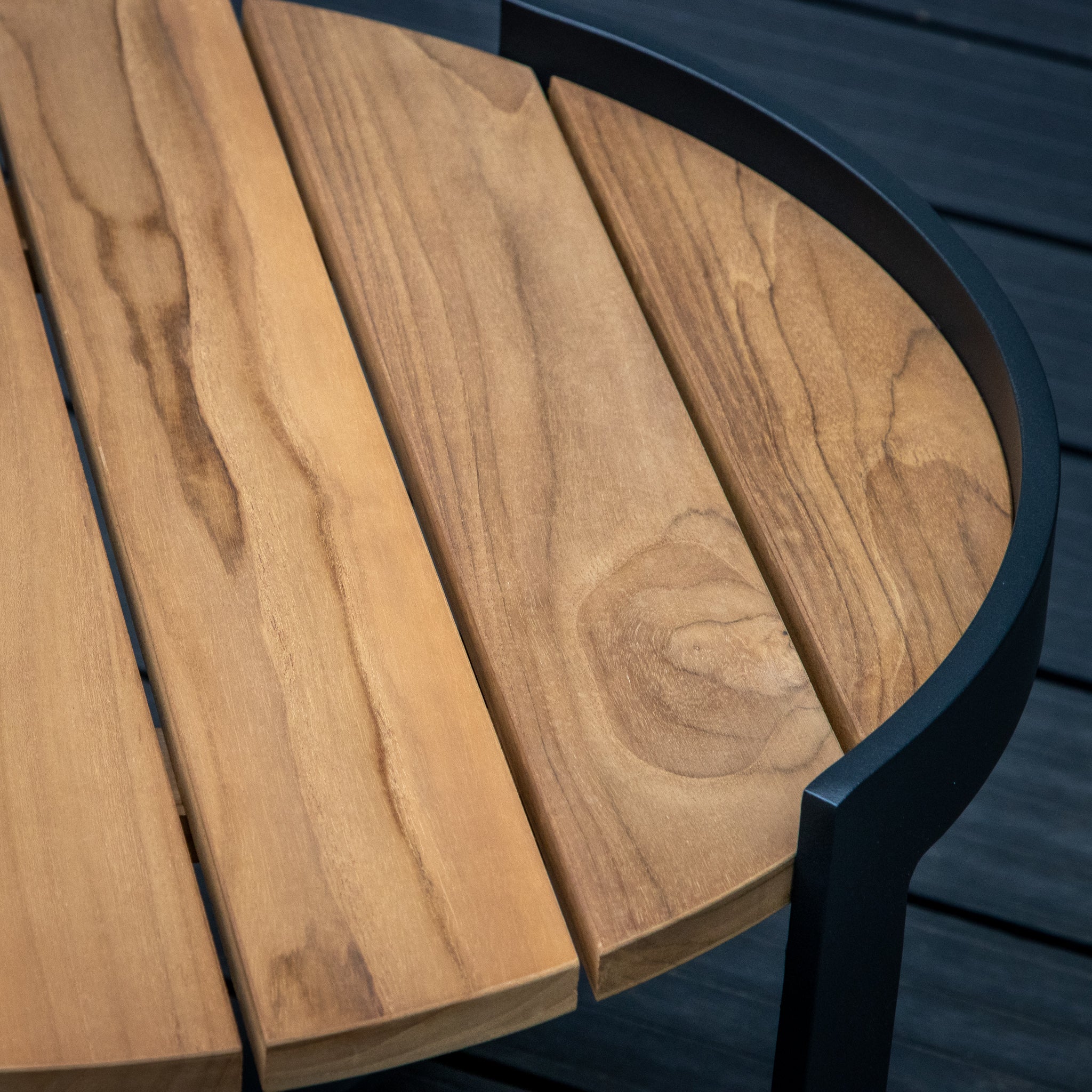 Close-up of a Bloom Coffee Table with Teak Top in Charcoal, featuring a wooden slatted teak top and a black metal frame, set on a dark wood deck background—perfect outdoor furniture.