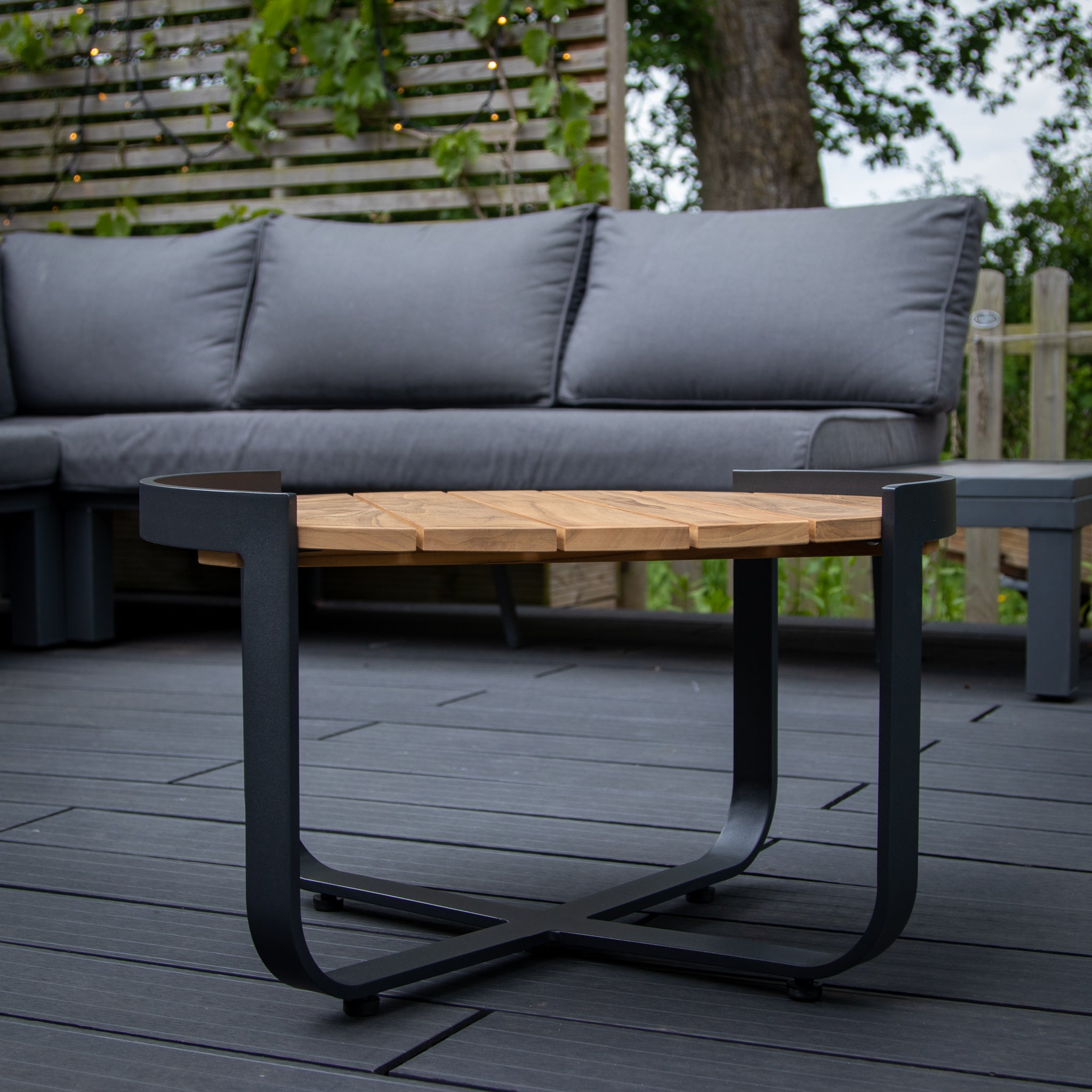 Modern outdoor furniture with a gray sofa and a Bloom Coffee Table with Teak Top in Charcoal on a dark deck, featuring a slatted privacy screen.