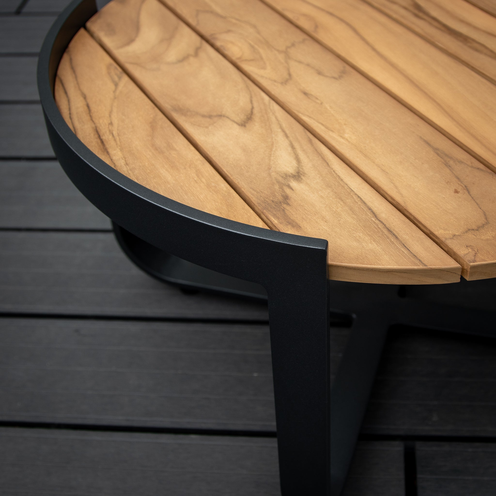 Close-up of the Bloom Coffee Table with Teak Top in Charcoal, featuring a round teak top and black metal legs on a dark wood floor.